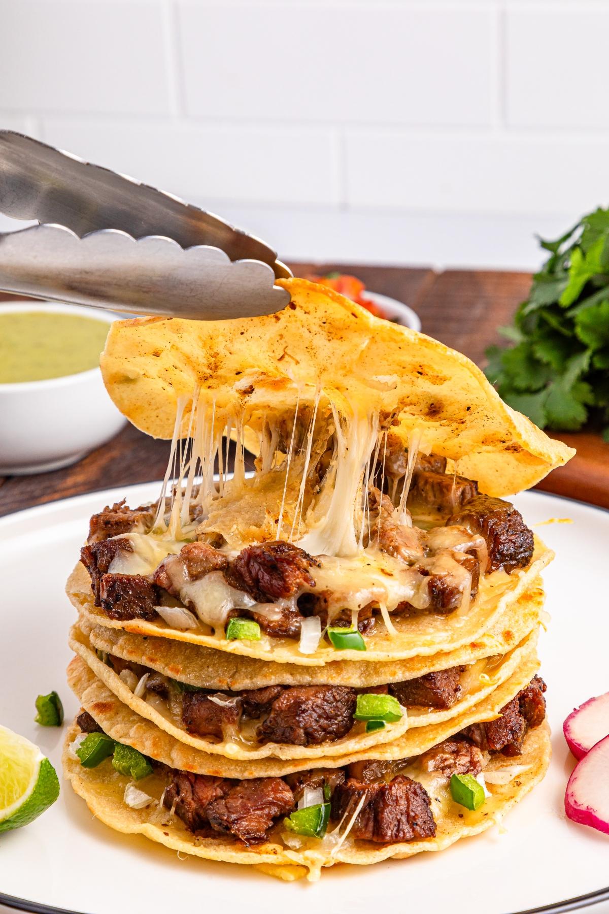 Metal tongs lift a cheesy beef taco from a stack of cheesy, meaty tacos on a white plate with garnishes.