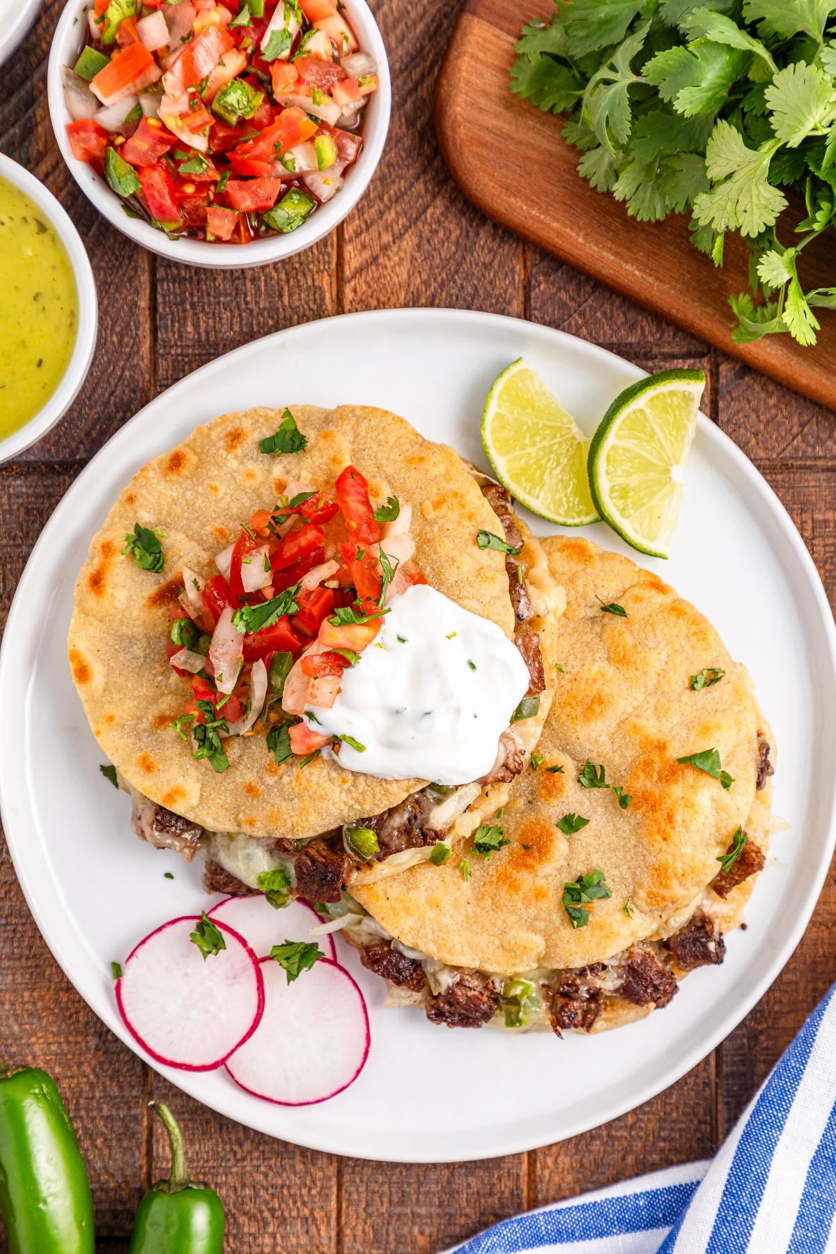 Two cheesy beef quesadillas topped with sour cream and salsa, served with lime, radish slices, and fresh cilantro.