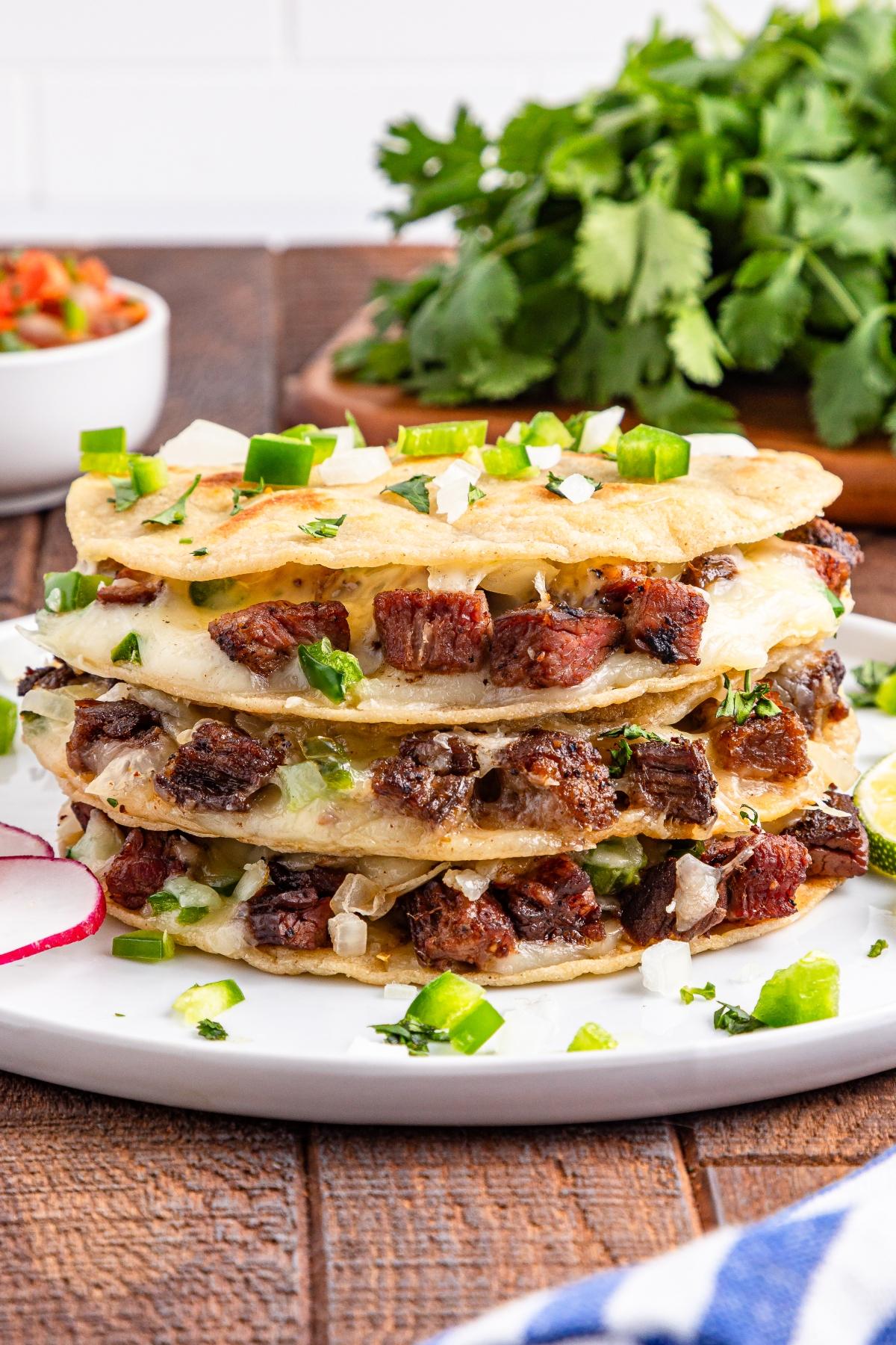 A stack of cheesy quesadillas filled with grilled beef, topped with chopped onions, cilantro, and jalape&ntilde;os.
