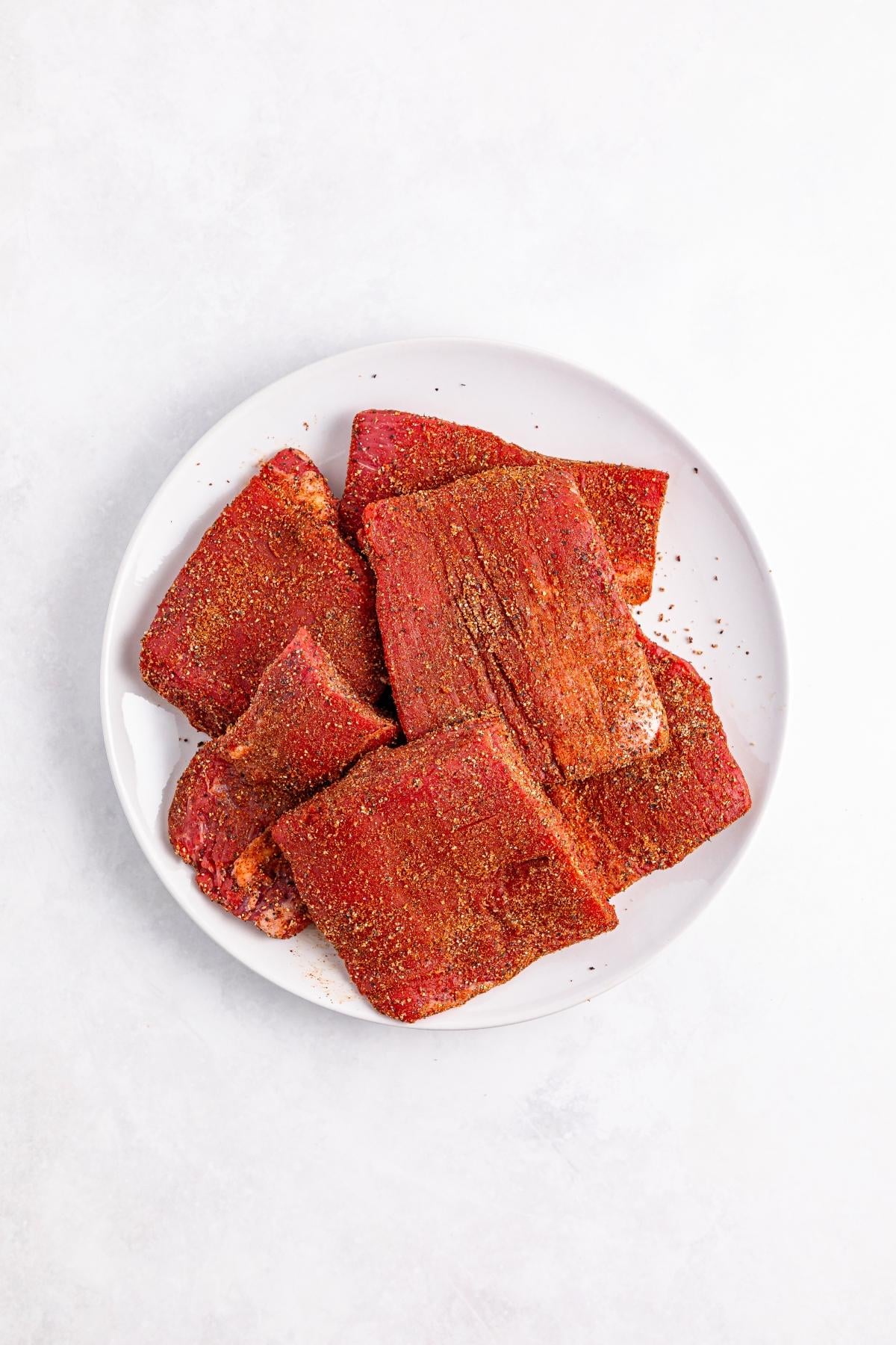 Raw, seasoned brisket slices arranged on a white plate against a light background.