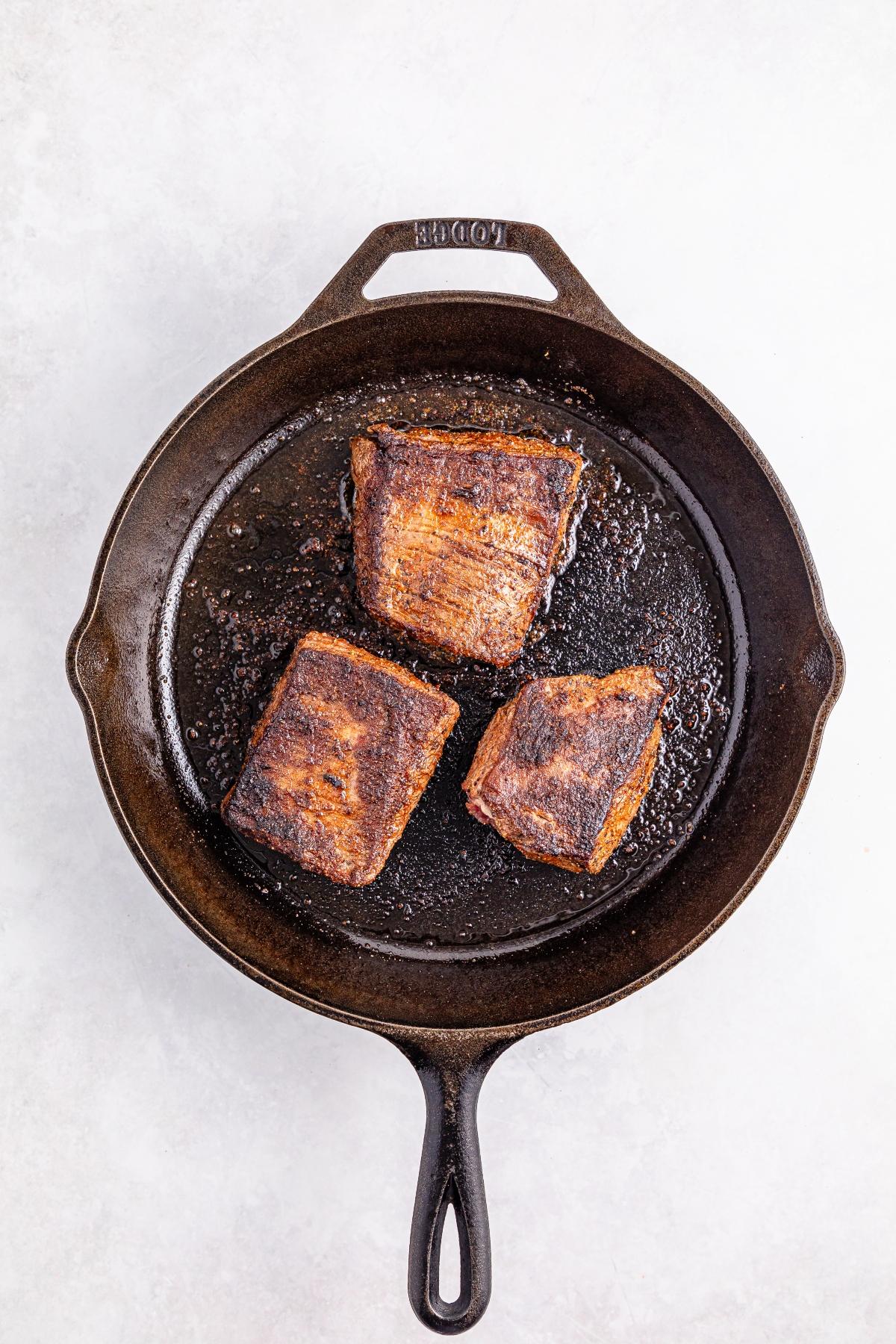 Three seared beef short ribs in a black cast iron skillet on a light background.
