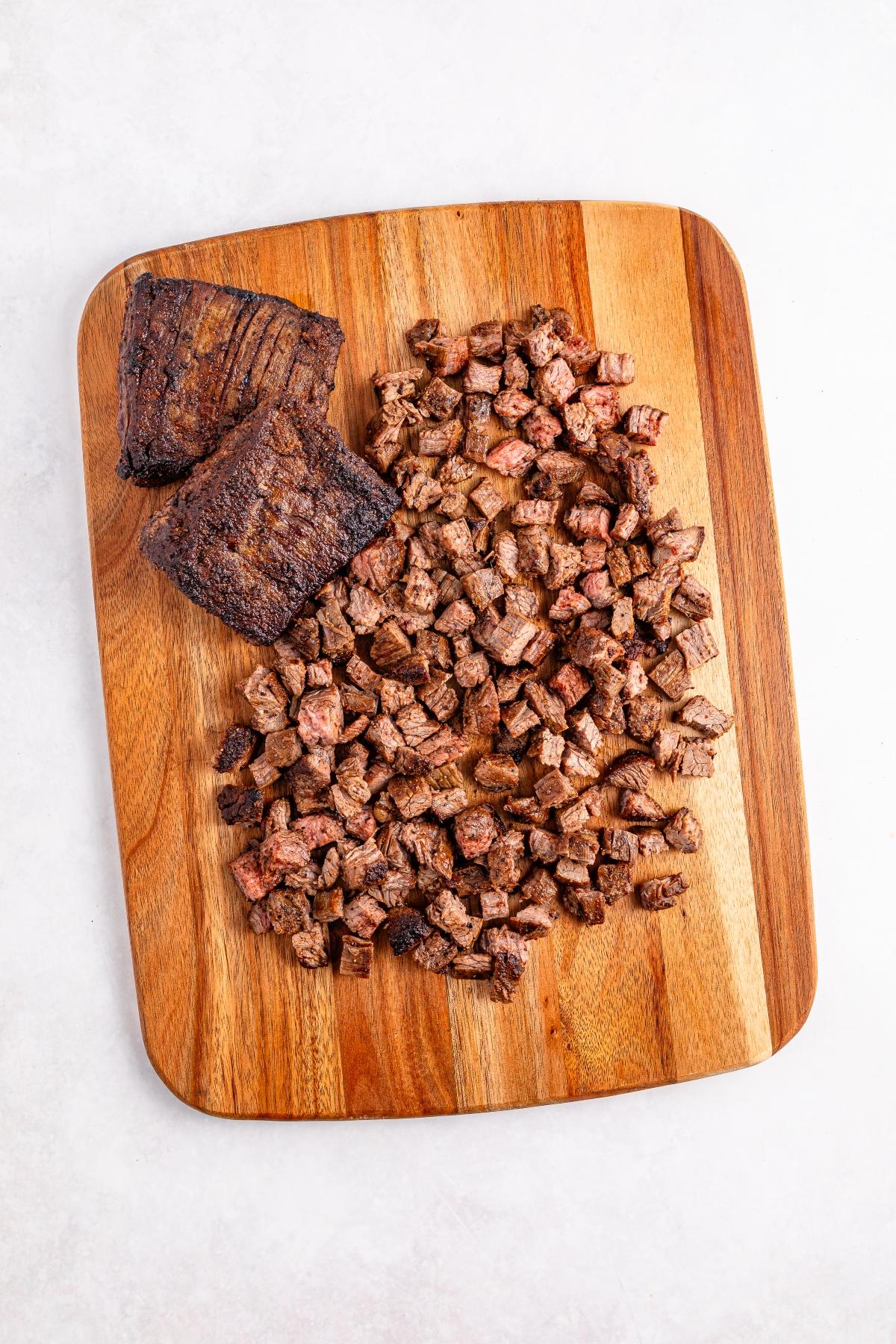Diced and whole cooked steak pieces on a wooden cutting board against a white background.