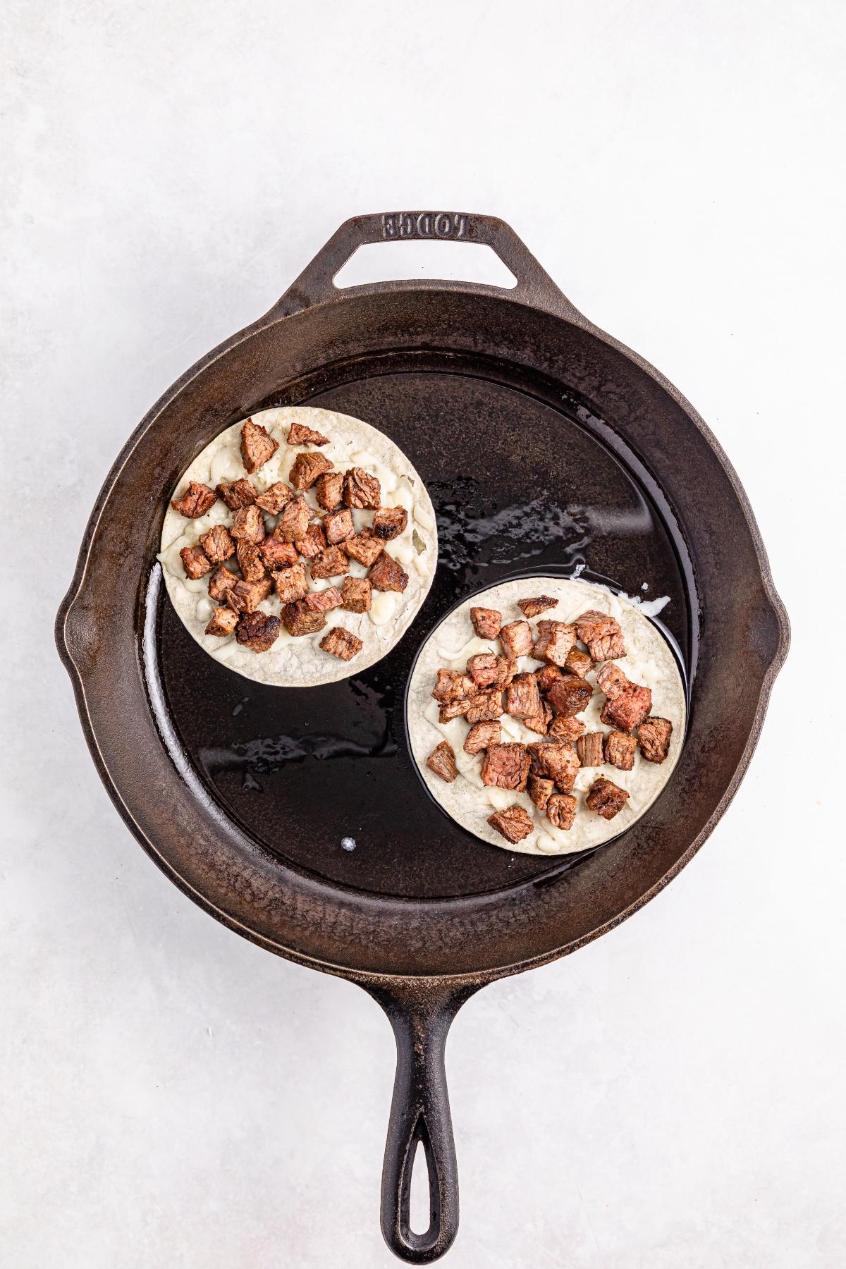 Two tortillas with diced cooked meat in a cast iron skillet on a white surface.