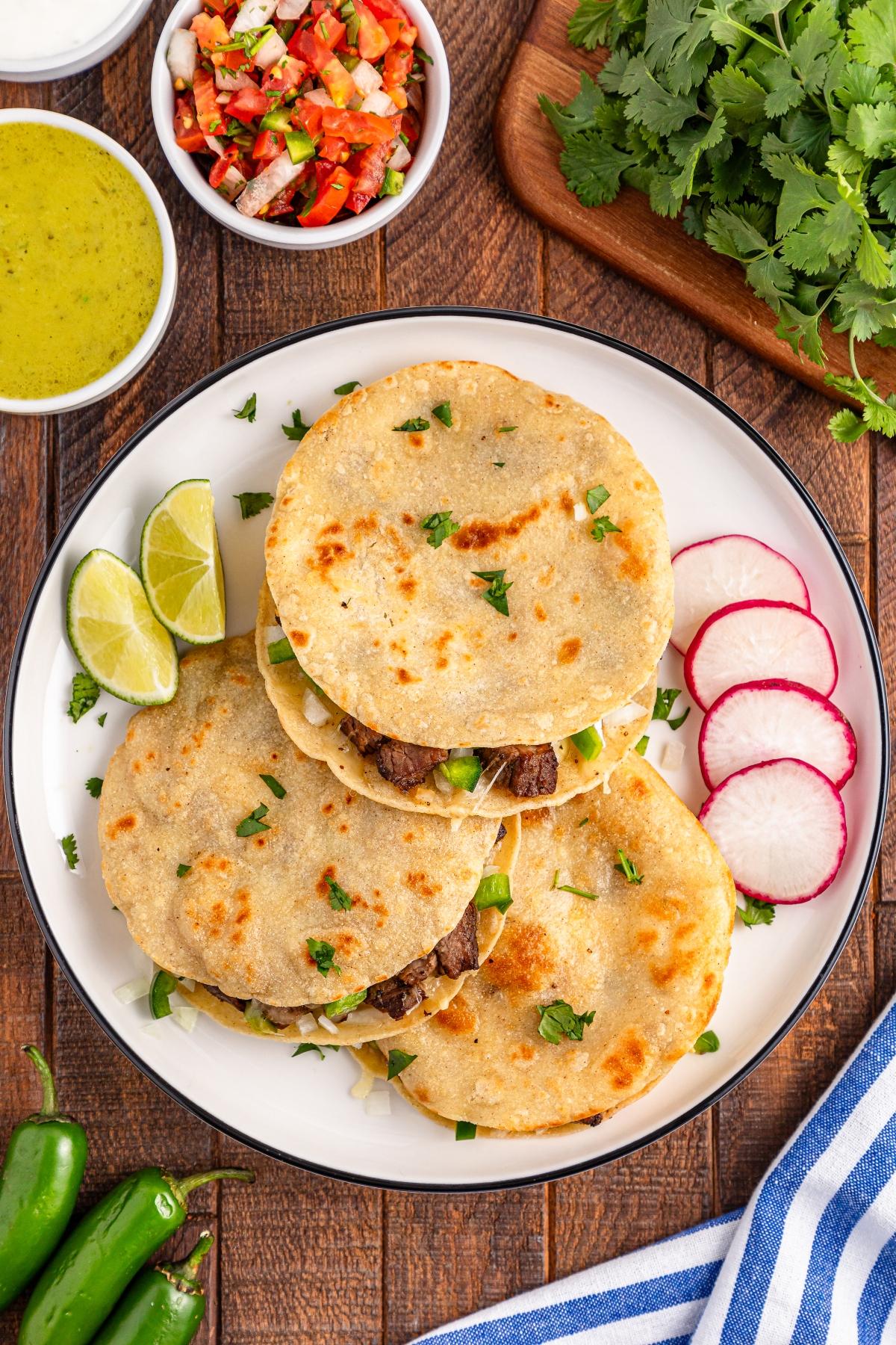 A plate of tacos with lime wedges, radish slices, salsa, and cilantro on a wooden table.