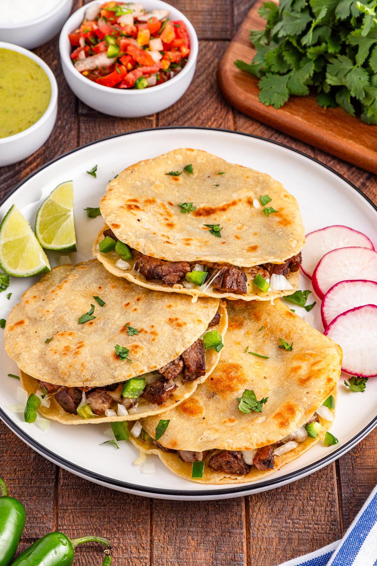 Three beef tacos with lime wedges and sliced radishes on a plate, with salsa and cilantro in the background.