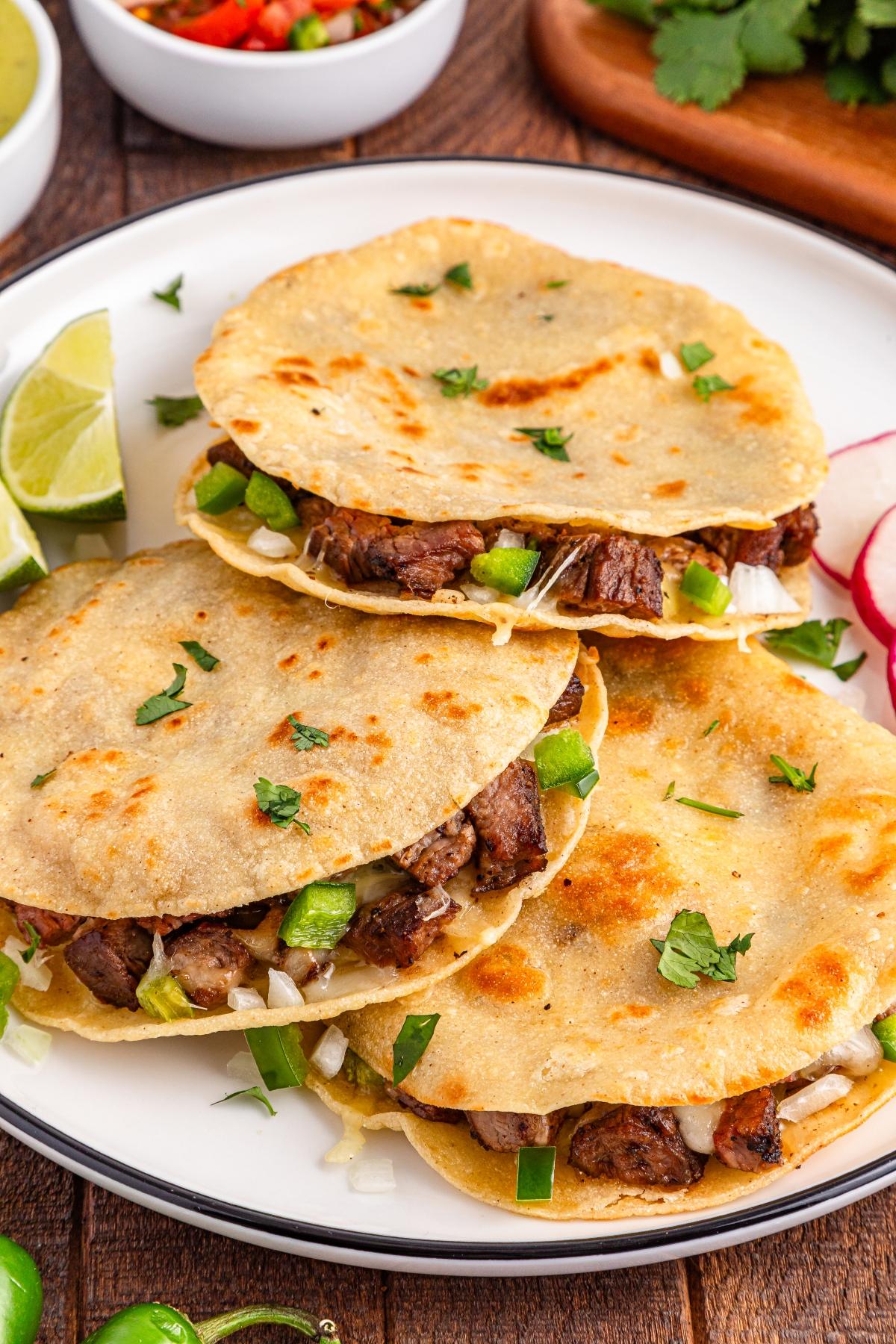 Three beef tacos with melted cheese, onions, and cilantro on a white plate, served with lime and radish slices.