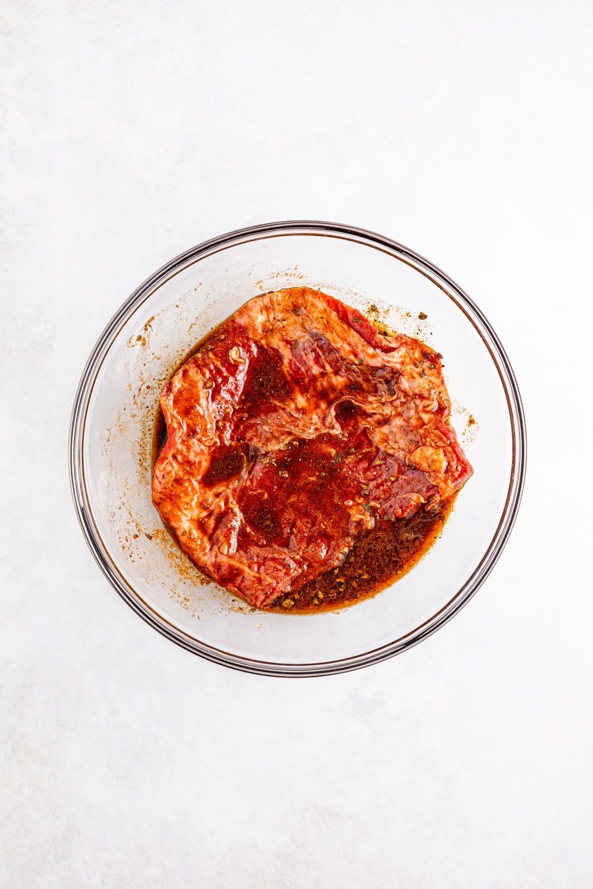Raw steak marinating in spices and sauce inside a clear glass bowl on a white surface.