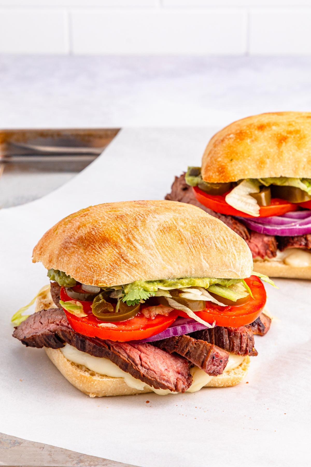 Two steak sandwiches with lettuce, tomato, red onion, and pickles on ciabatta rolls, on parchment paper.