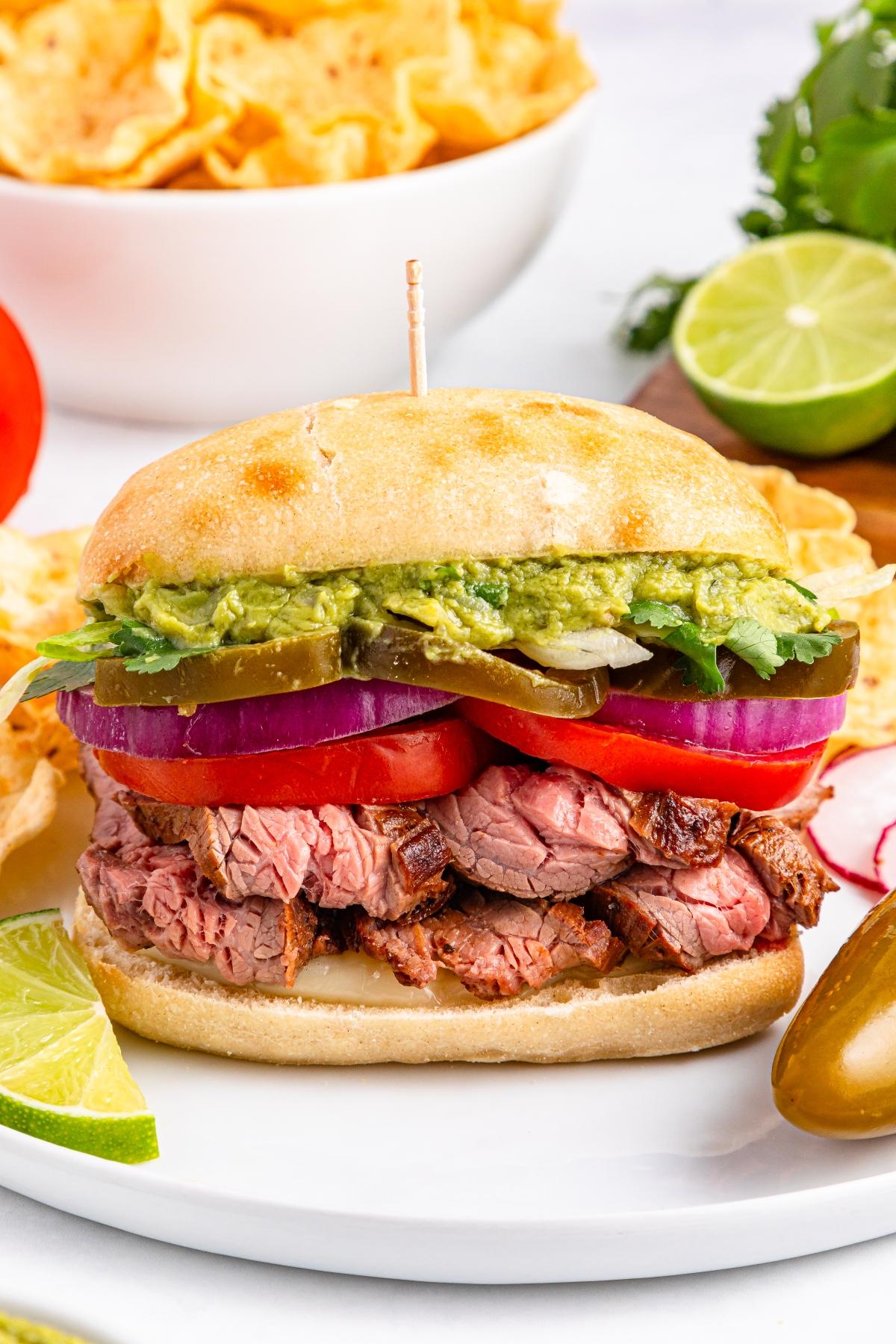 A steak sandwich with tomato, onion, lettuce, jalape&ntilde;os, and guacamole on a plate with lime and chips.