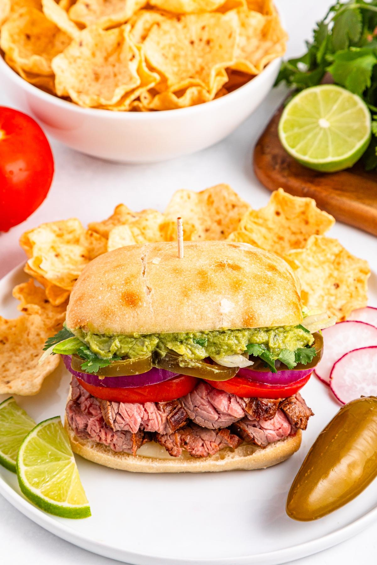 A steak sandwich with guacamole, tomato, red onion, and chips on a plate with lime wedges and a jalape&ntilde;o.