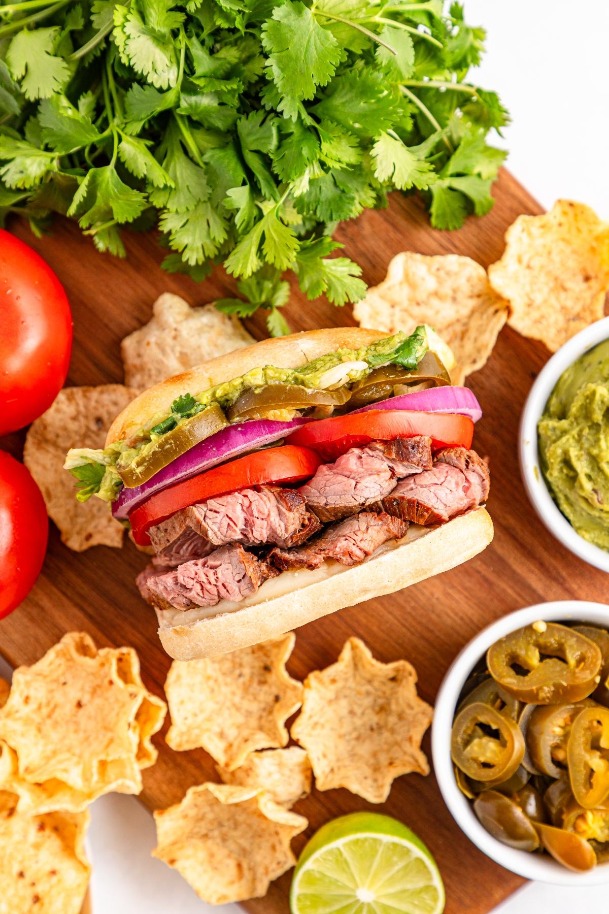 A steak sandwich with tomato, onion, and avocado, surrounded by chips, jalape&ntilde;os, guacamole, and cilantro.