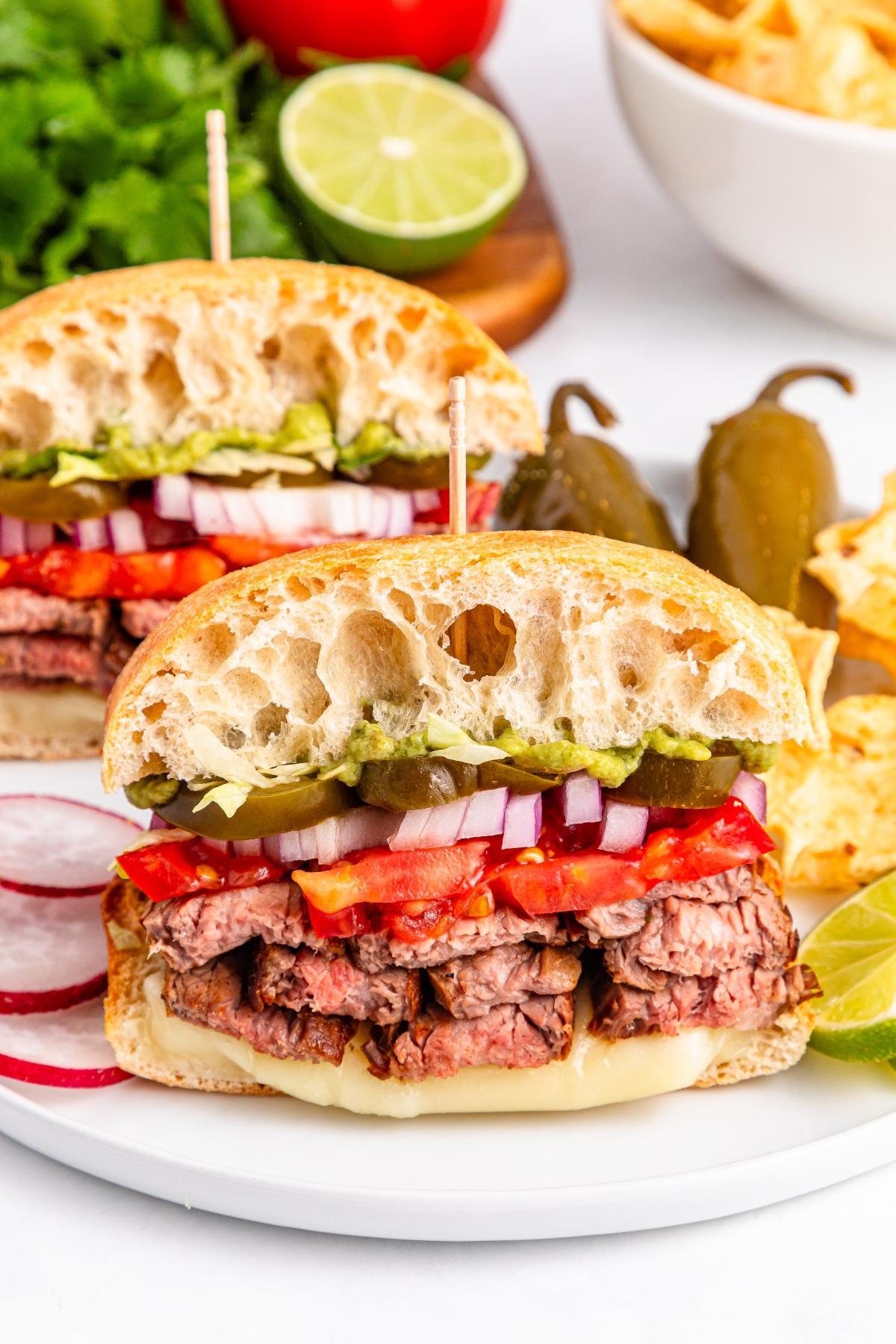 A steak sandwich with onions, tomatoes, pickles, and cheese on ciabatta bread, served with chips and lime.