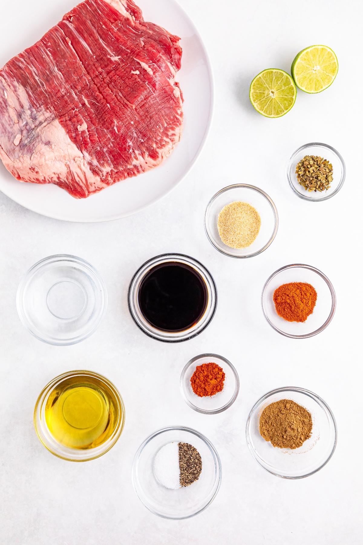 Raw flank steak on a plate with lime halves and small bowls of spices, oil, and soy sauce on a white surface.