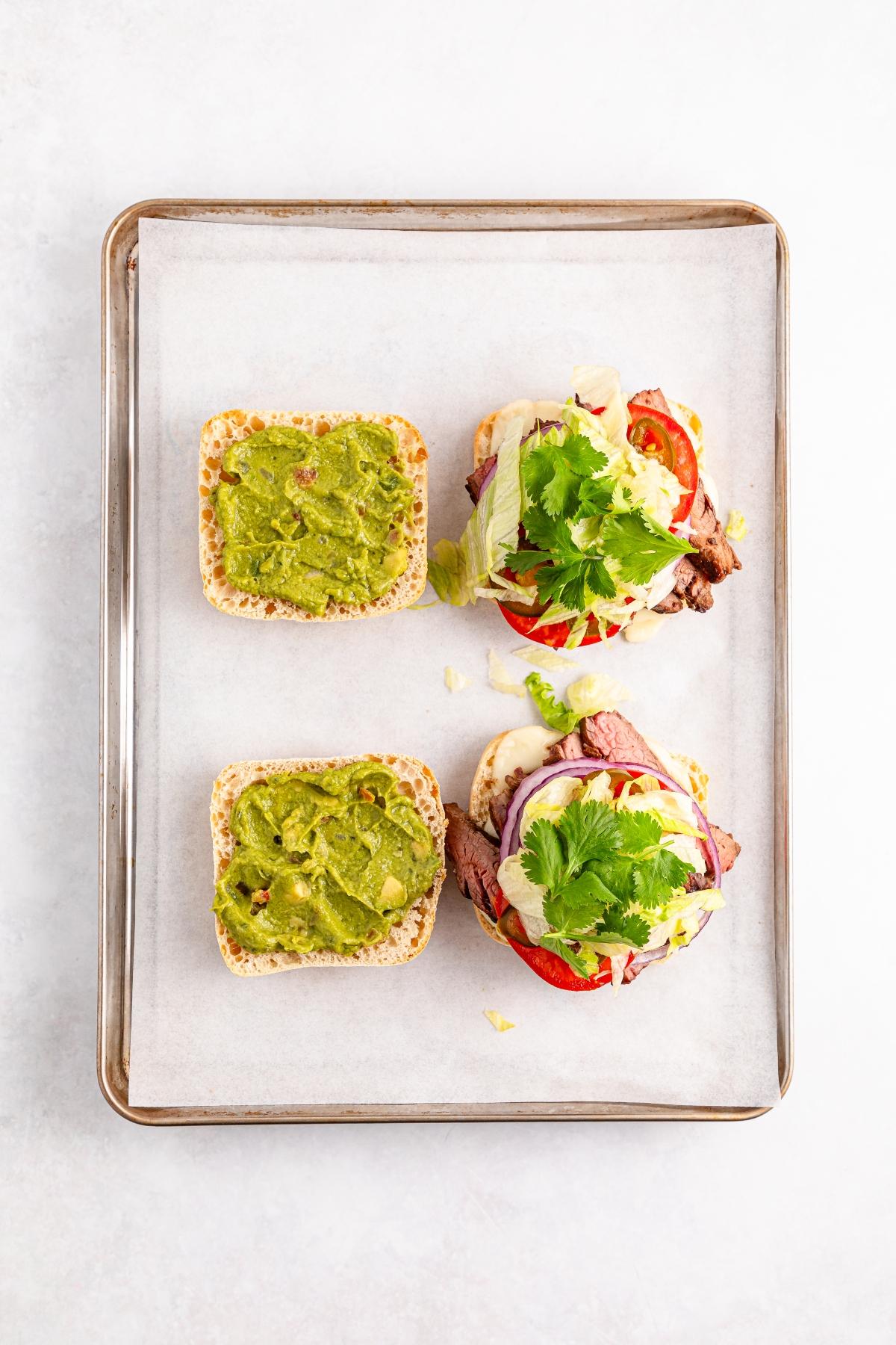 Two open steak sandwiches with guacamole, tomato, onion, and cilantro on a baking tray lined with parchment paper.