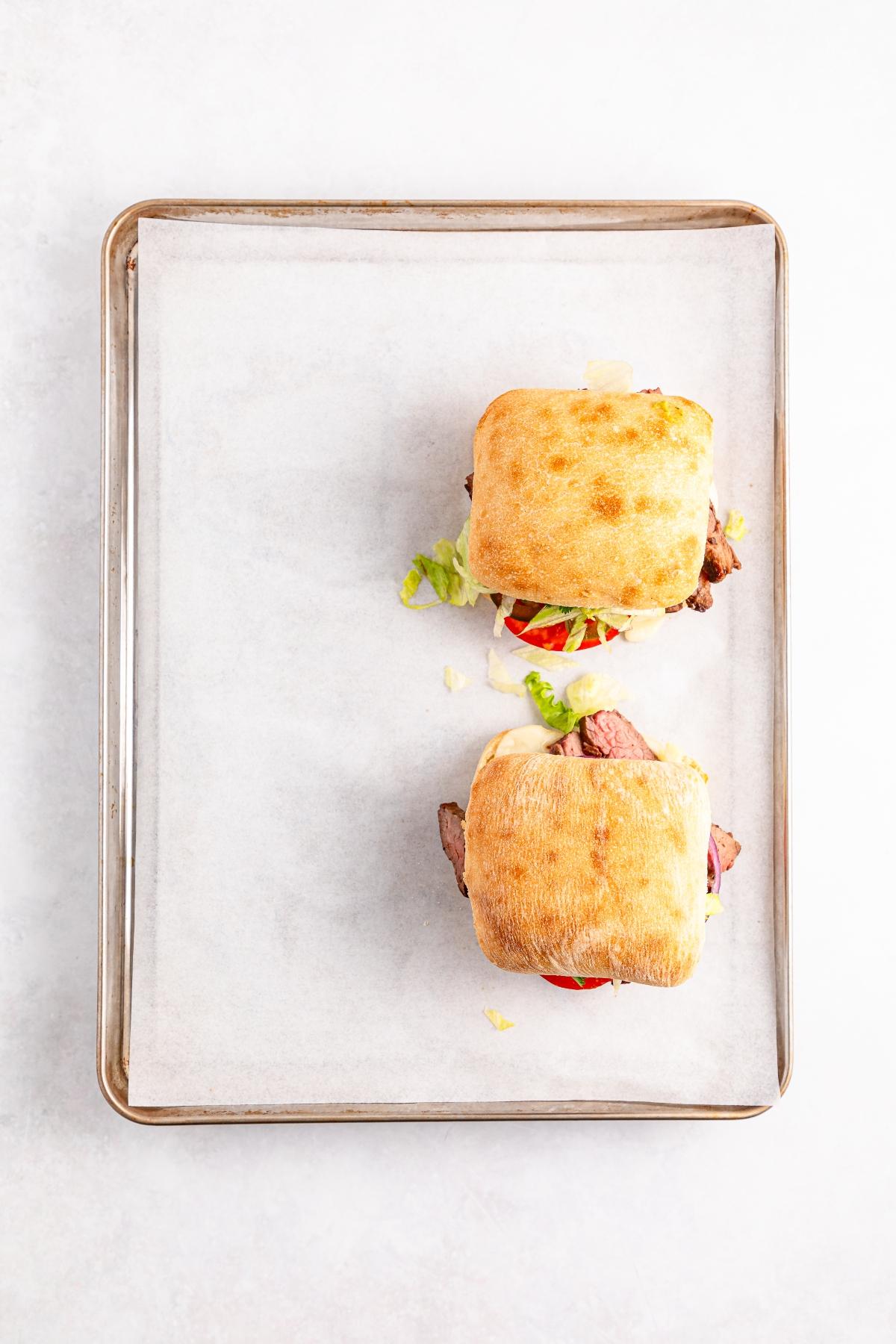 Two ciabatta sandwiches with visible lettuce and tomato on a parchment-lined baking tray.