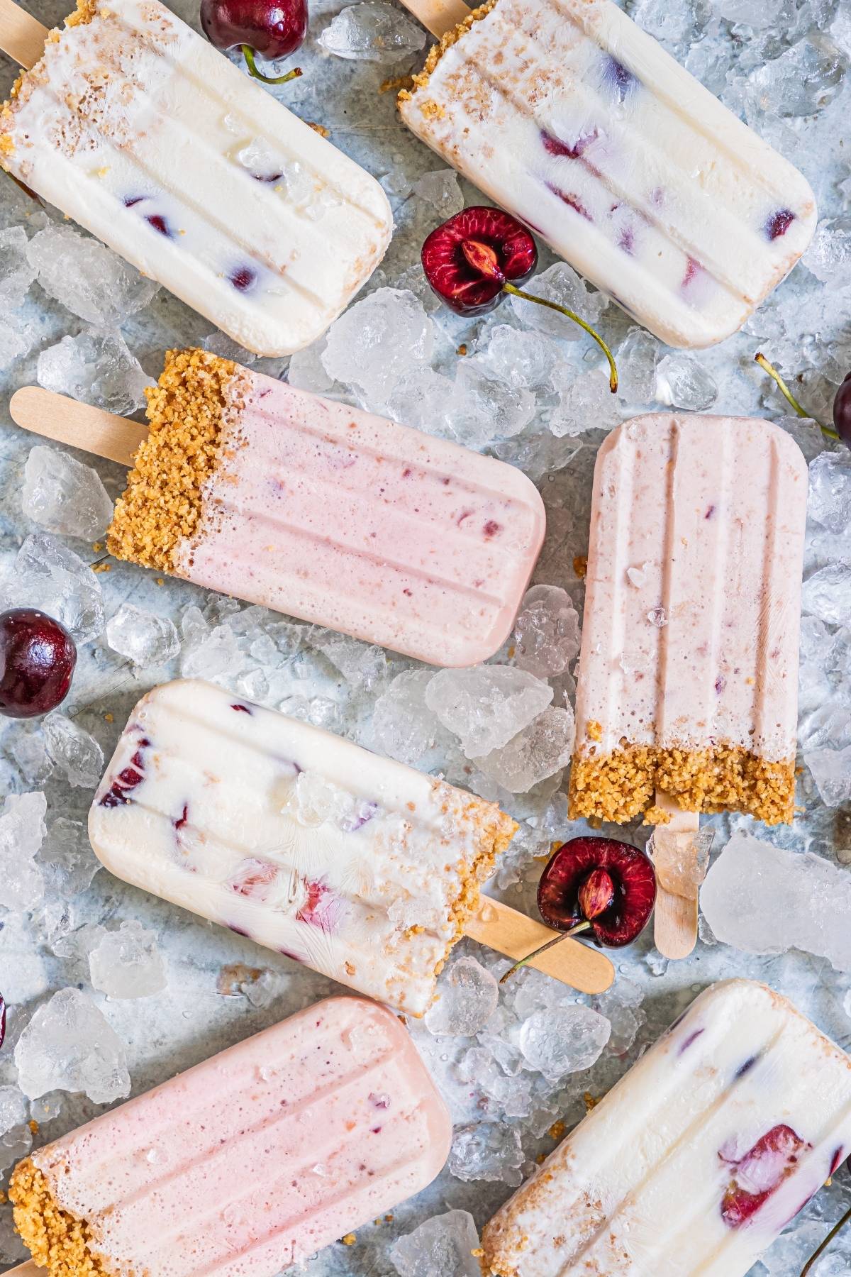 Cherry Cheesecake Popsicles with graham cracker crust on ice, surrounded by fresh cherries—a perfect summer treat recipe for anyone craving a cool dessert.