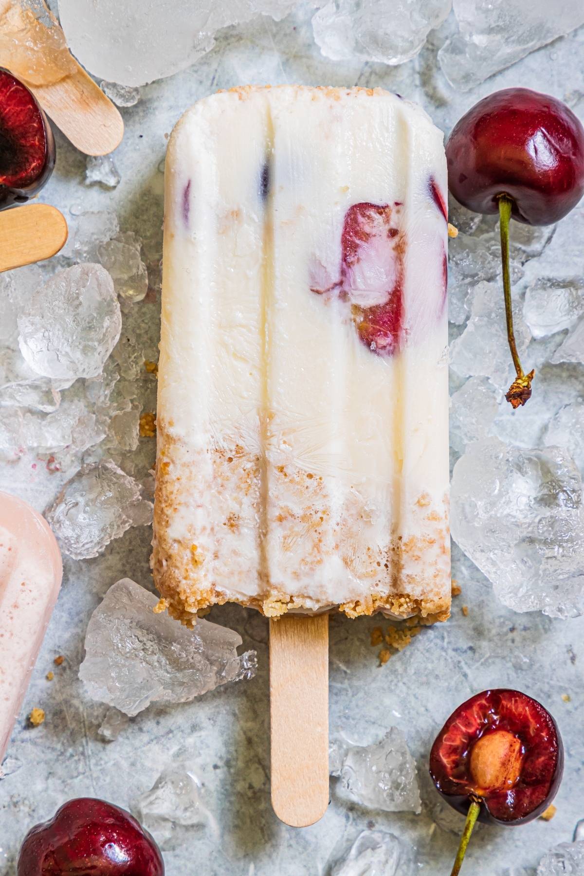 A creamy Cherry Cheesecake Popsicle with a bite taken out, surrounded by ice and fresh cherries—the perfect summer treat recipe.