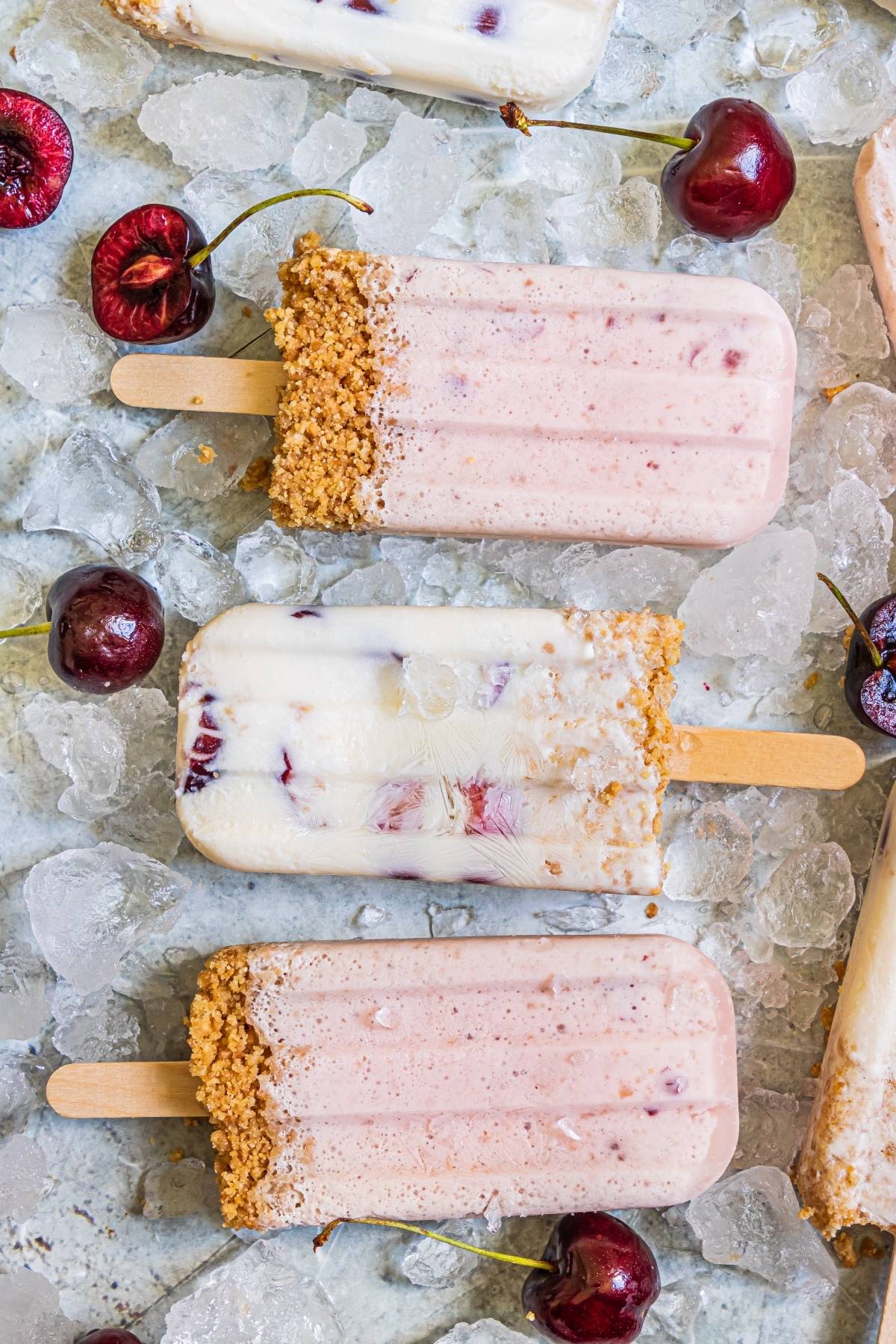 Three cherry cheesecake popsicles with graham cracker crusts on ice, surrounded by fresh cherries—a perfect cherry dessert popsicle and summer treat recipe.