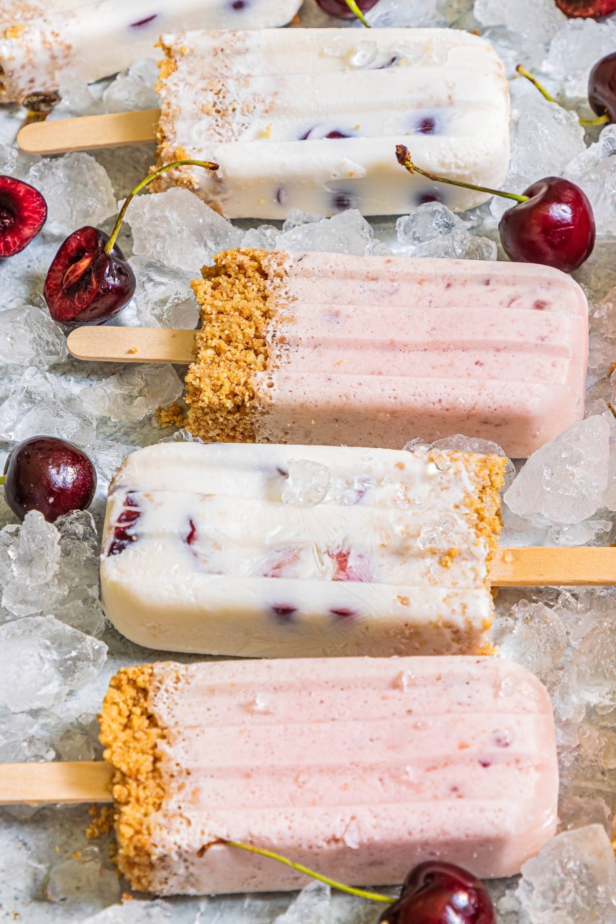 Assorted Cherry Cheesecake Popsicles on ice, with fresh cherries and a graham cracker crumb coating—this creamy popsicle recipe is the ultimate cherry dessert popsicle for summer.