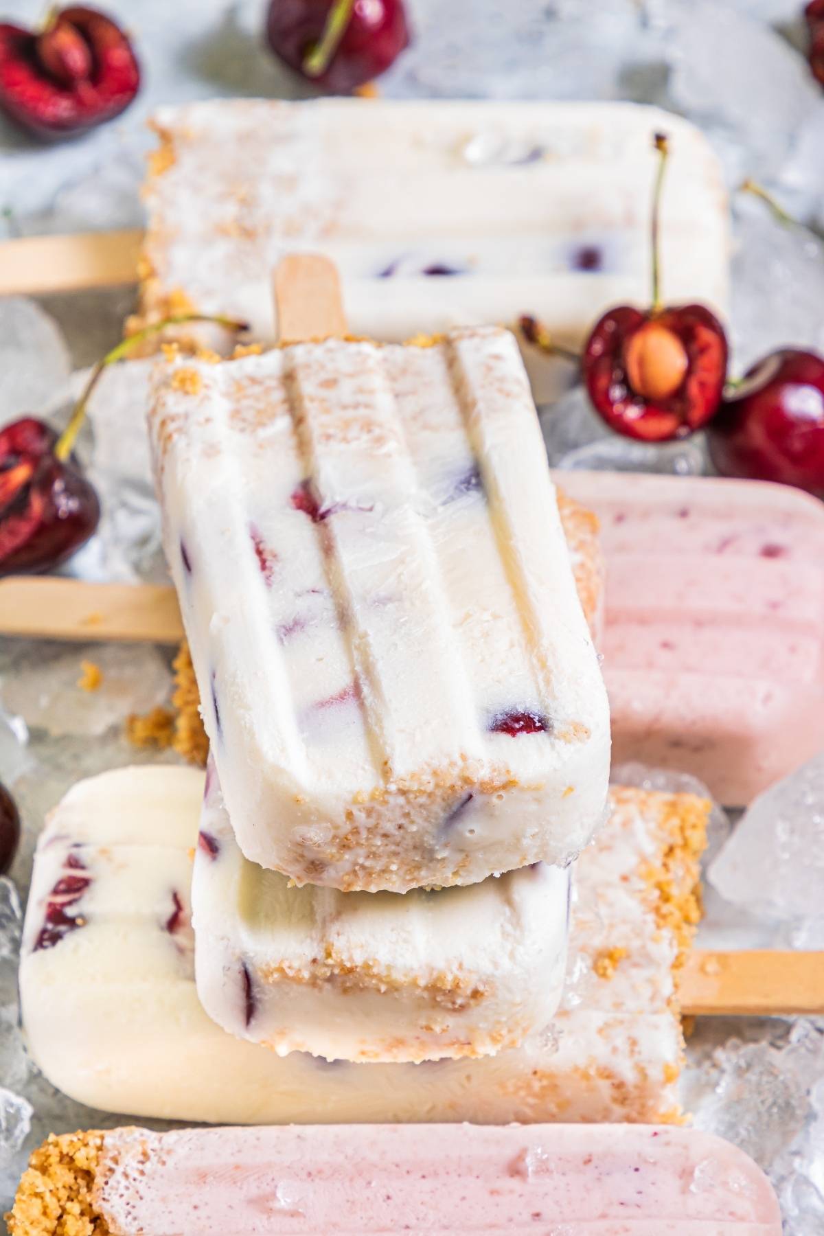 Stacked cherry cheesecake popsicles with graham cracker crusts on ice, surrounded by fresh cherries—an irresistible cherry dessert popsicle and a perfect recipe with fresh cherries for summer.
