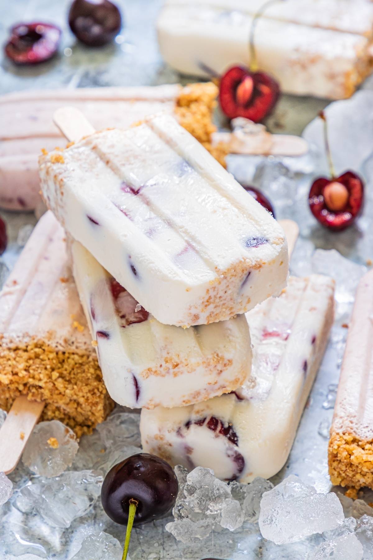 Three creamy popsicles made with a cherry dessert popsicle twist and graham cracker crumbs, stacked on ice with fresh cherries—a perfect summer treat recipe.