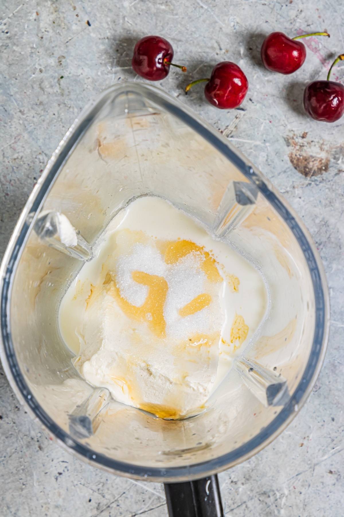 Blender with milk, sugar, flour, and eggs inside; three cherries are on the countertop nearby—perfect for a summer treat recipe or a delicious recipe with fresh cherries.