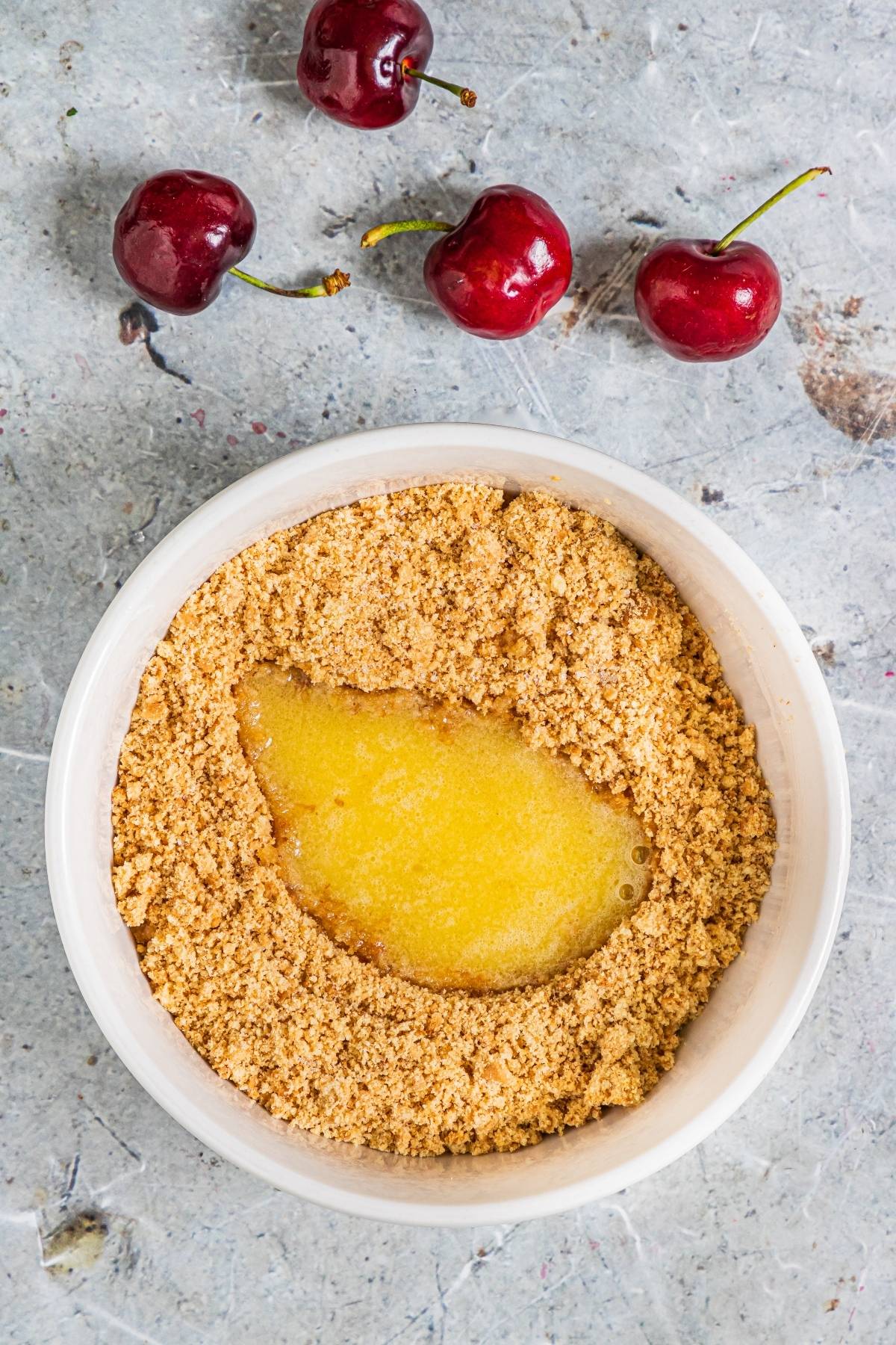 A bowl of graham cracker crumbs and melted butter beside three fresh cherries on a gray surface—perfect inspiration for a creamy popsicle recipe like Cherry Cheesecake Popsicles.