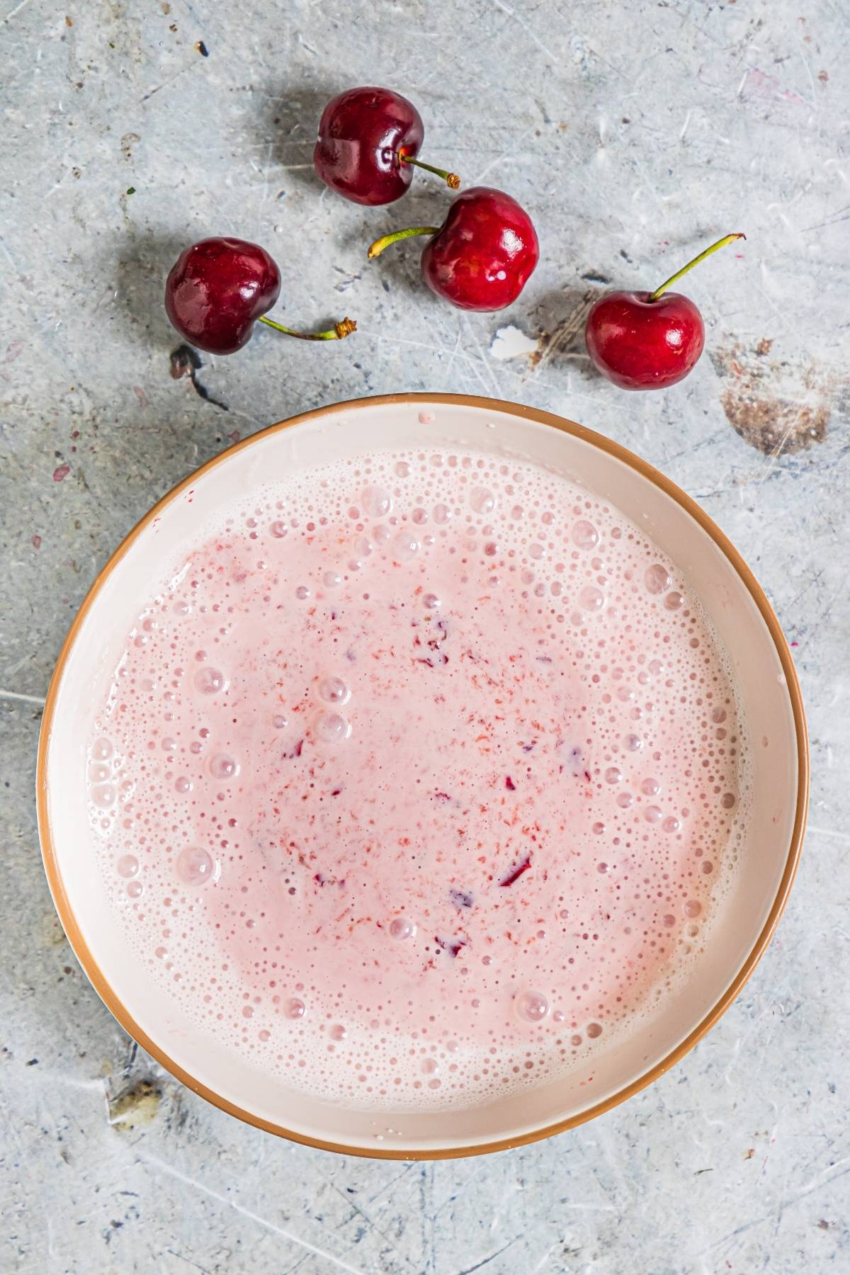 A bowl of pink cherry smoothie sits on a gray surface beside three fresh cherries, perfect inspiration for a creamy popsicle recipe or a refreshing summer treat recipe.