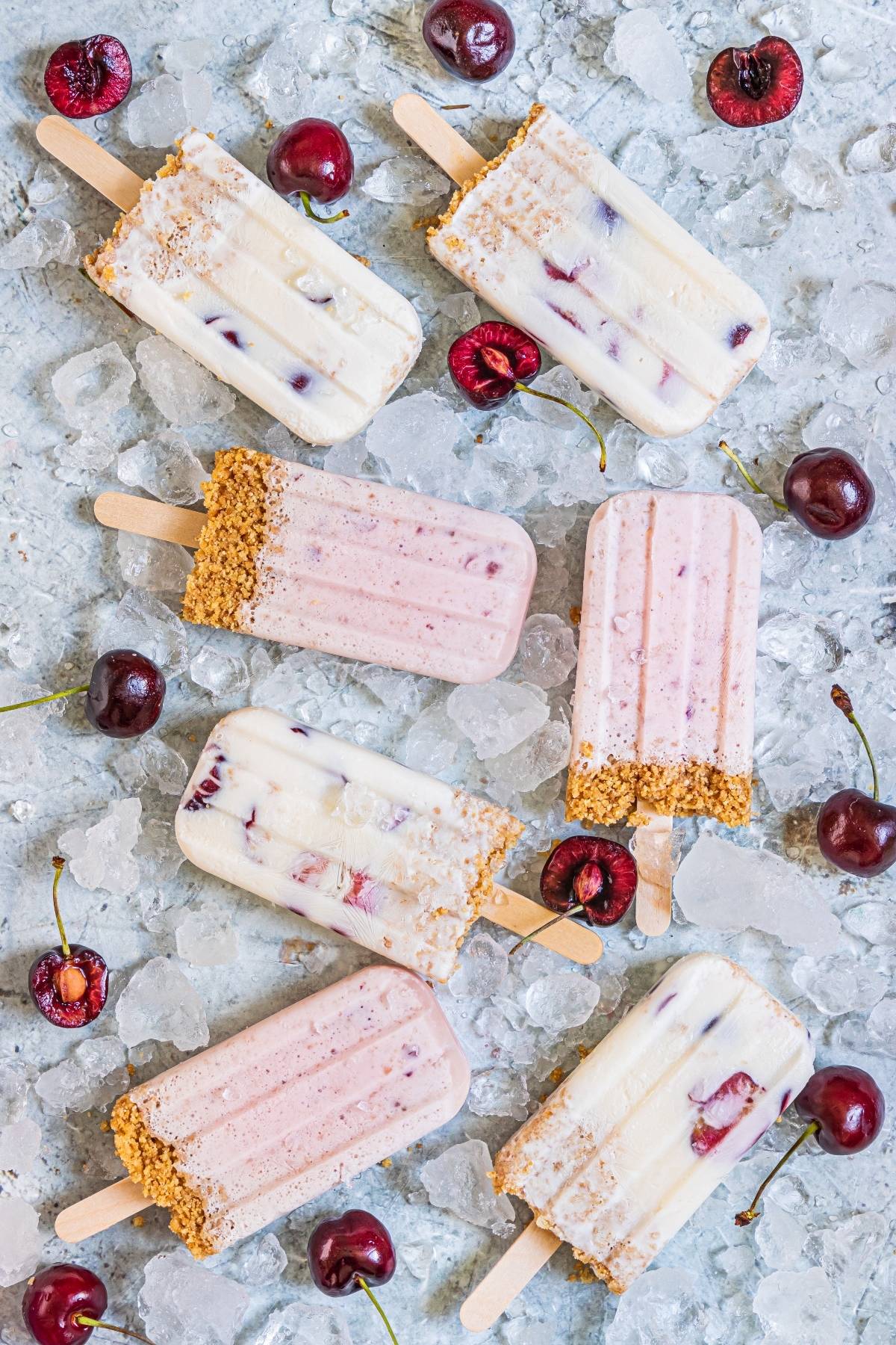 Assorted cherry cheesecake popsicles with graham cracker crust on ice, surrounded by fresh cherries—a creamy popsicle recipe perfect for any cherry dessert popsicle lover.