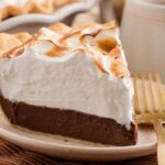 A decadent slice of chocolate meringue pie rests on a plate with a gold fork, while the remaining chocolate meringue pie sits elegantly in the background.