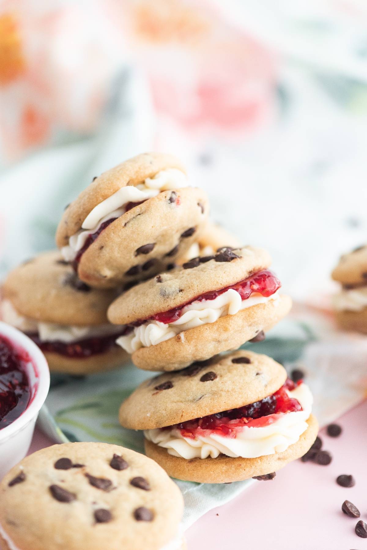 Stacked chocolate chip cookie sandwiches filled with creamy sandwich cookie filling and red jam, on a pastel floral cloth—truly some of the best sandwich cookies with jam.