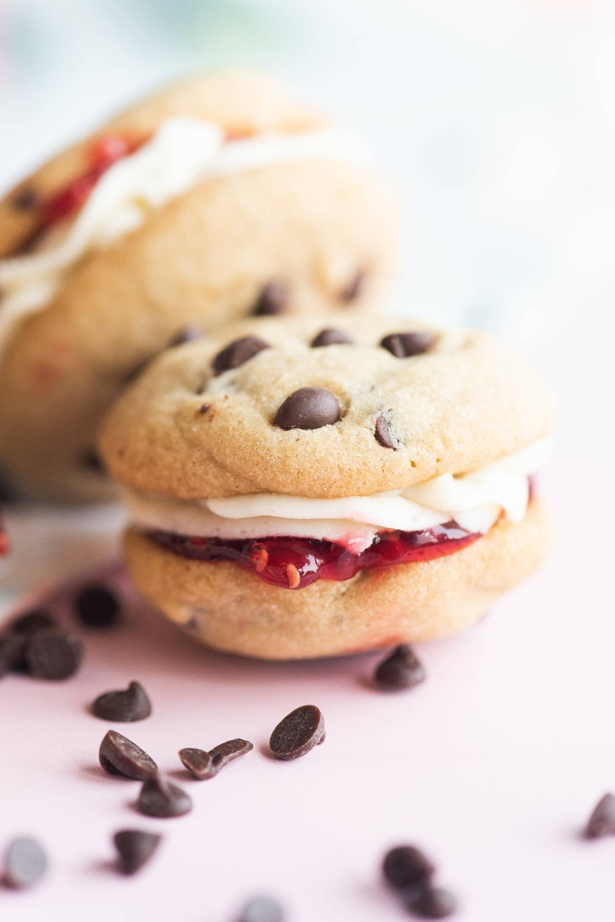Two chocolate chip sandwich cookies with cream and jam filling, surrounded by chocolate chips—perfect for fans of the best sandwich cookies with jam.