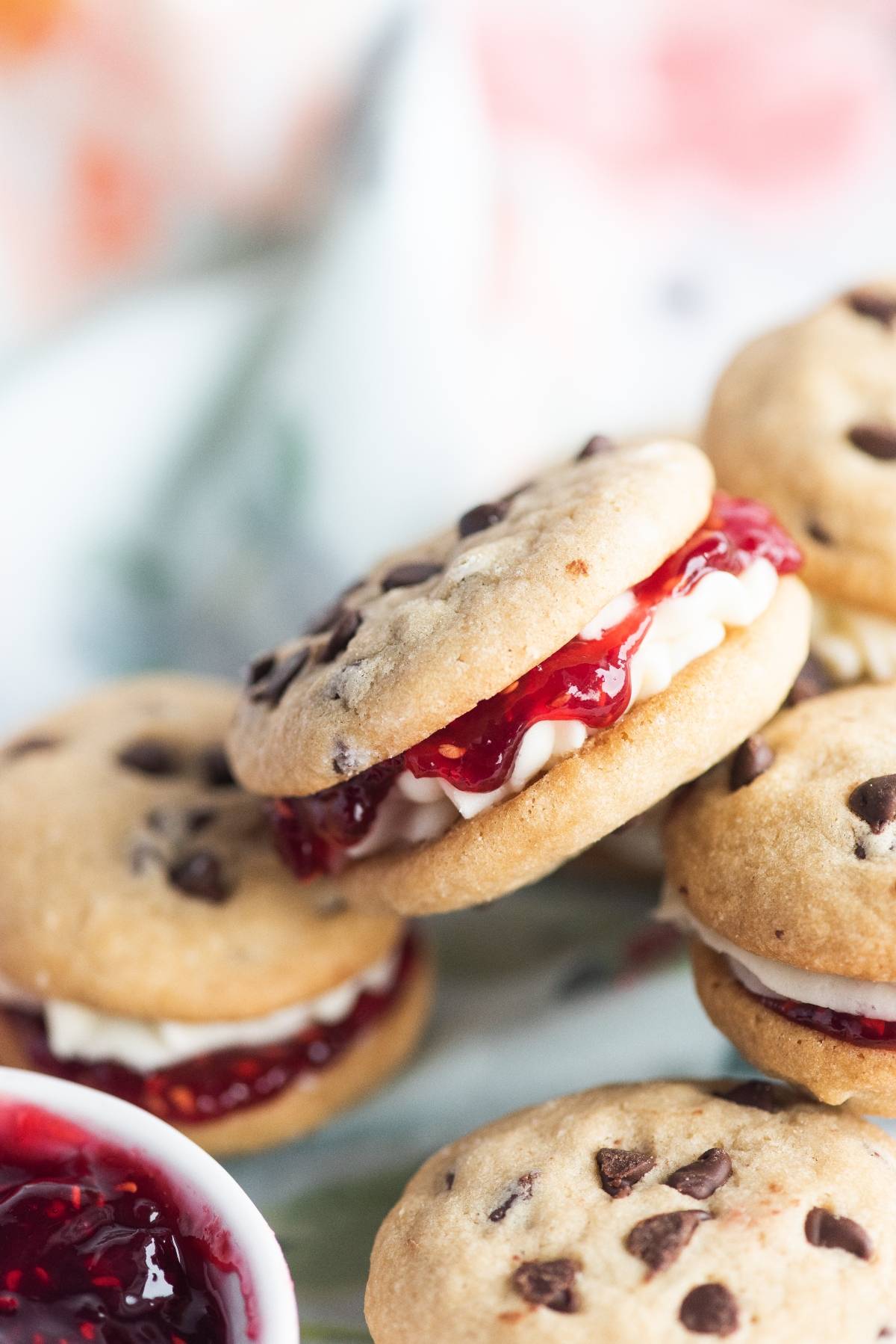 Chocolate chip cookie sandwiches with the best filling for sandwich cookies—rich cream and berry jam—are stacked on a light surface.