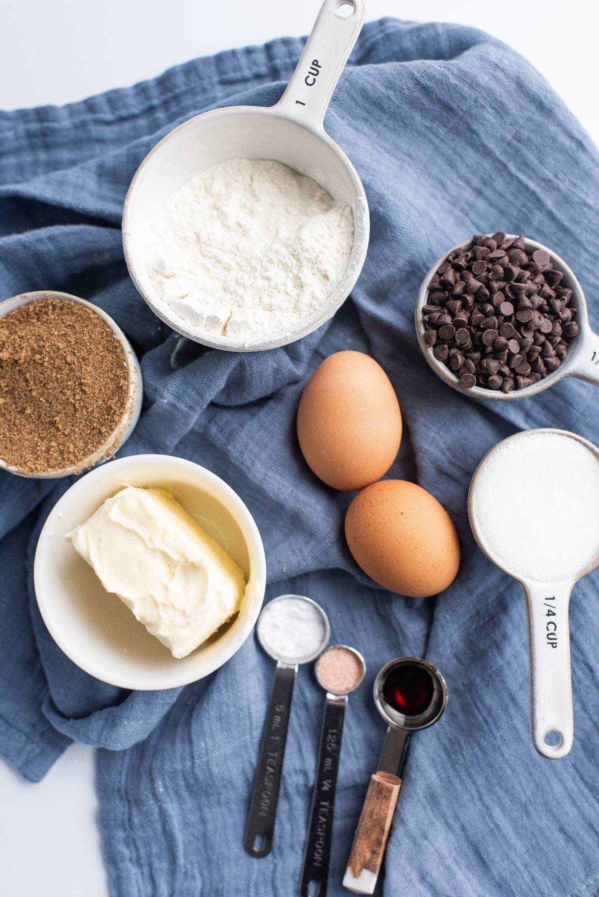 Baking ingredients on a blue cloth: flour, eggs, butter, brown sugar, white sugar, chocolate chips, and measuring spoons—everything you need to create the best sandwich cookies with jam or decadent Chocolate Chip Buttercream Sandwiches.