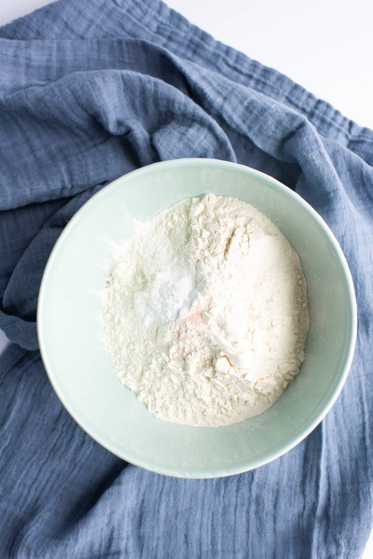 A bowl of flour and baking powder on a blue cloth, ready to help you craft the best sandwich cookies with jam.