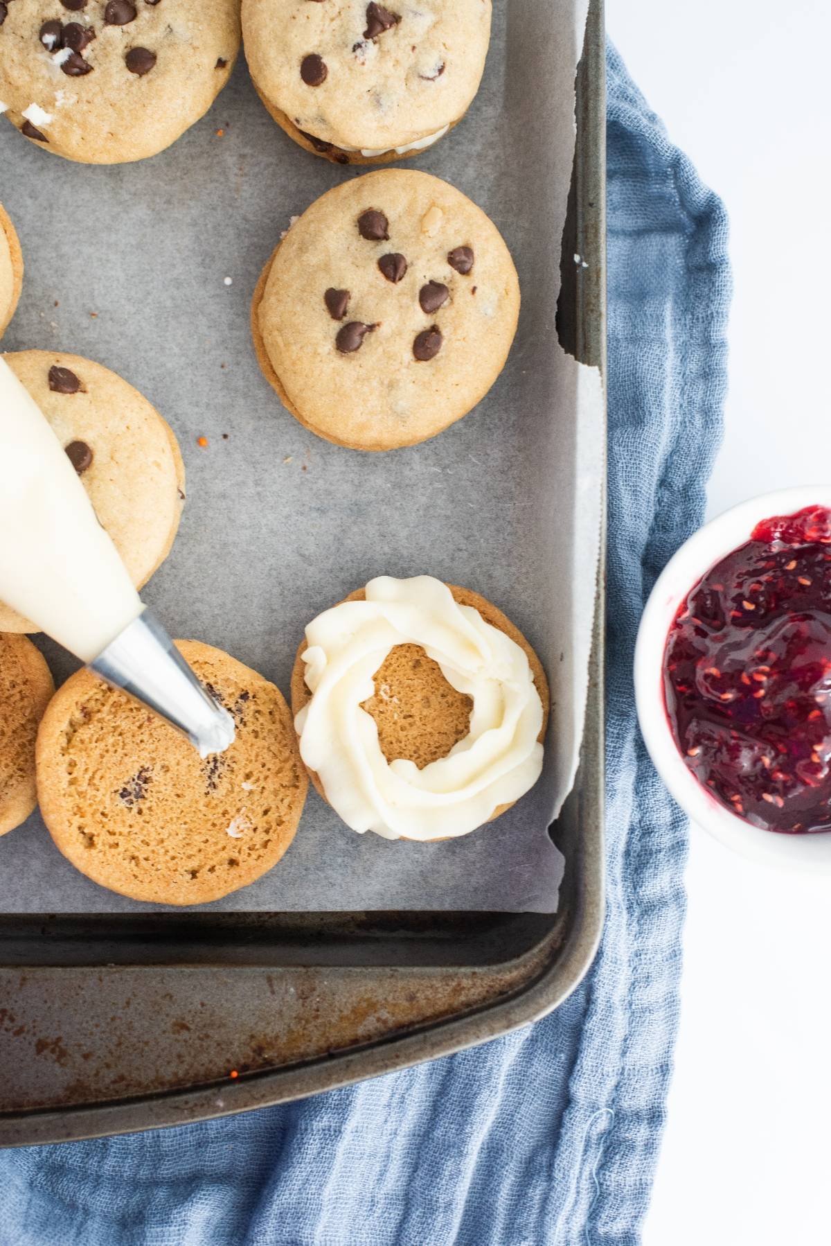 A piping bag adds frosting to a cookie; more cookies and a bowl of berry jam are nearby on a blue cloth—perfect inspiration for the best filling for sandwich cookies like Chocolate Chip Buttercream Sandwiches.