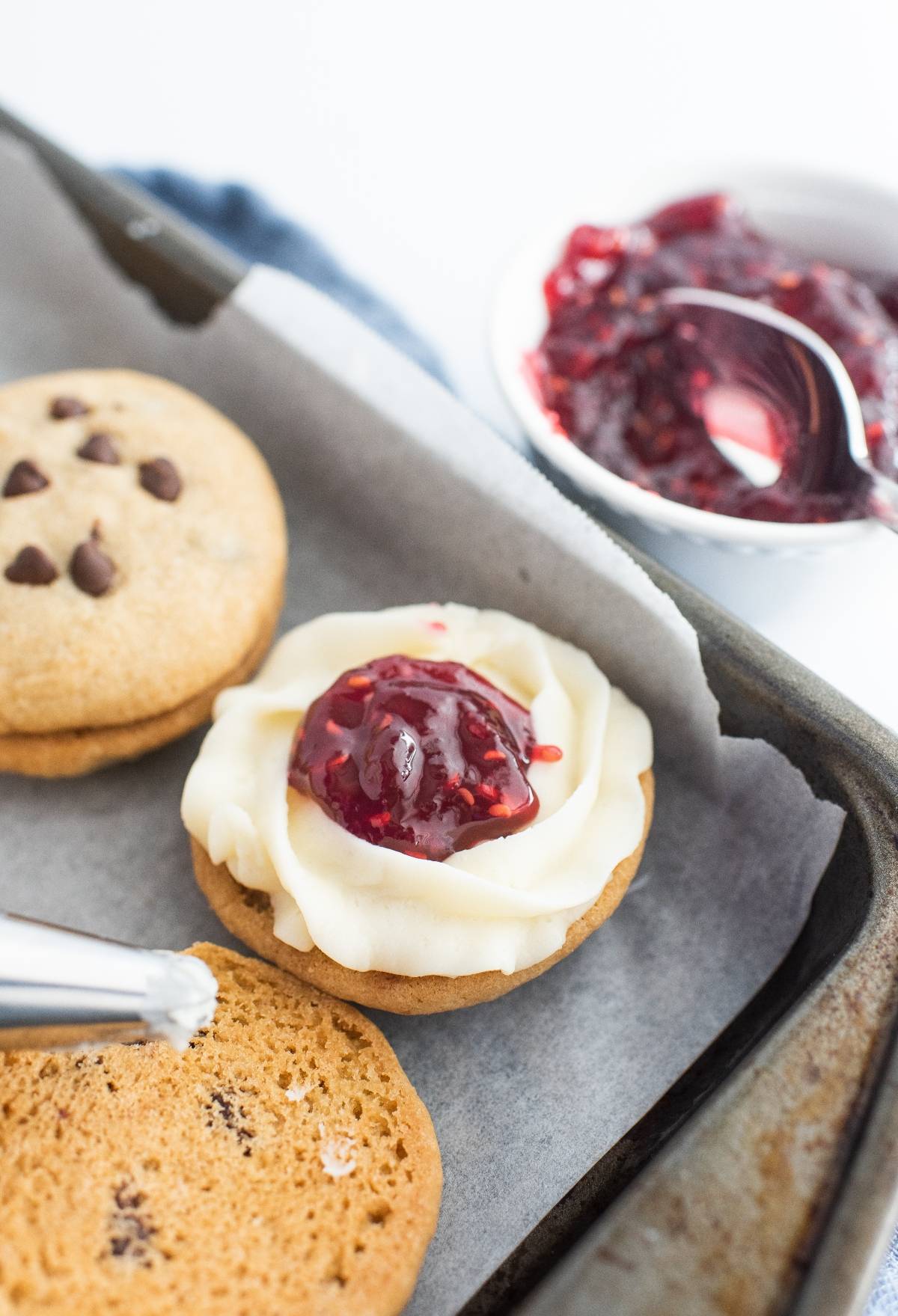 A cookie topped with white frosting and raspberry jam on a baking tray, showcasing the best sandwich cookies with jam, with extra jam and cookies nearby.