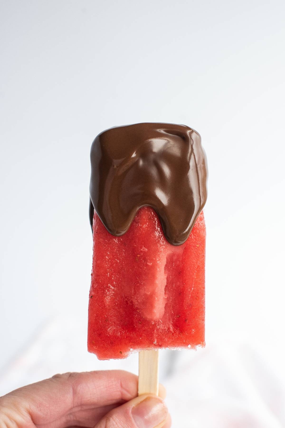 Hand holding a red popsicle partially dipped in melted chocolate against a plain white background—a tempting Chocolate Covered Strawberry Popsicles idea and perfect easy homemade popsicles summer treat recipe.