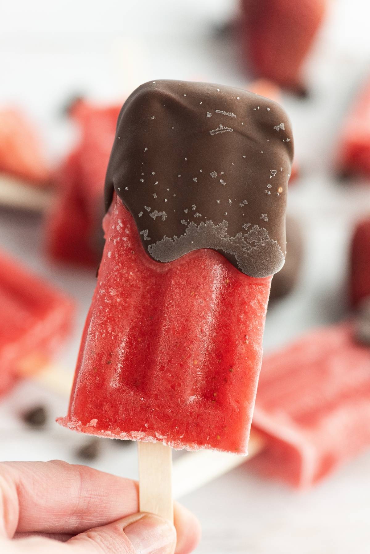 Hand holding a red popsicle dipped in chocolate—perfect for anyone craving Chocolate Covered Strawberry Popsicles. More easy homemade popsicles are blurred in the background, making this an irresistible dipped fruit popsicle recipe.