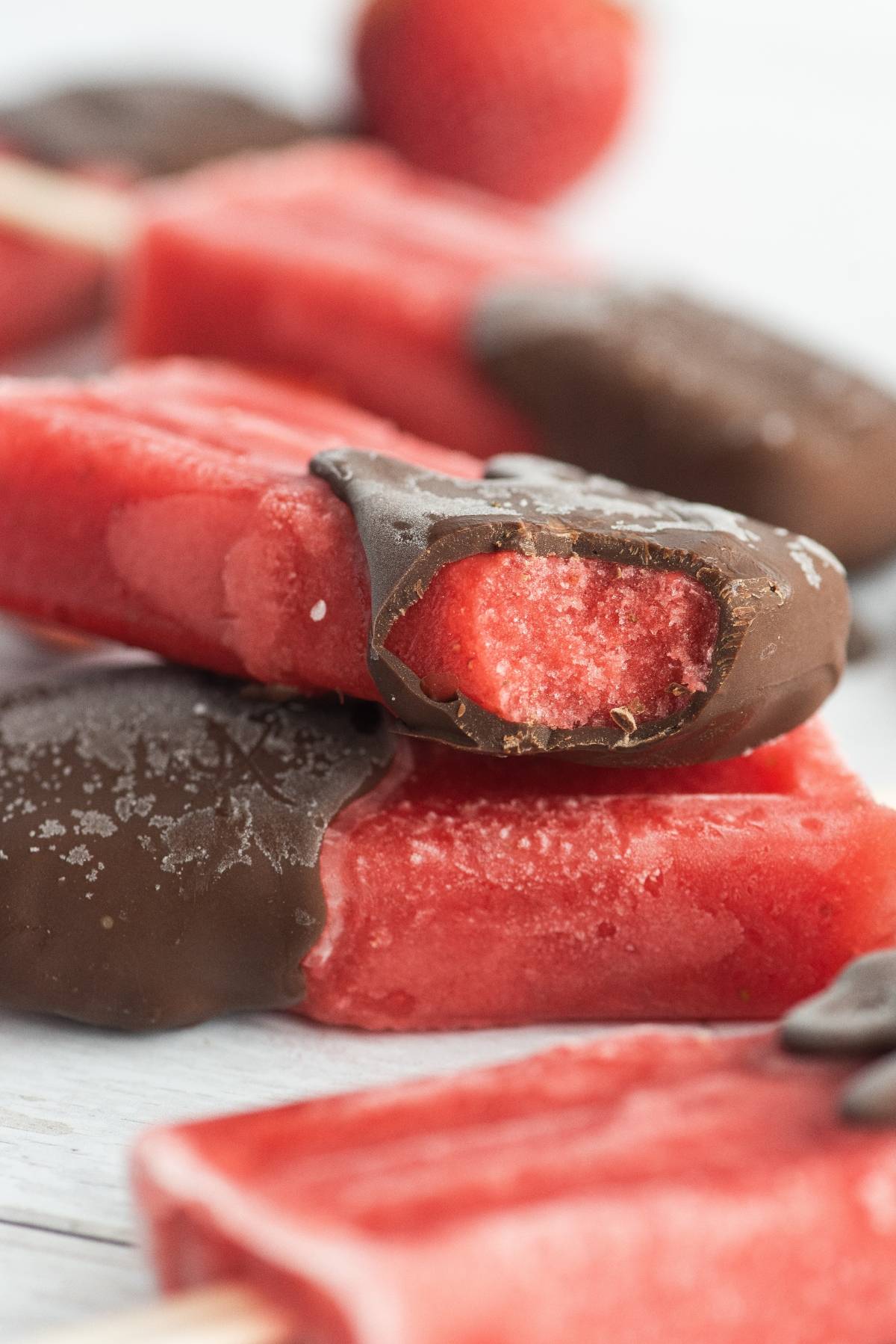 Chocolate Covered Strawberry Popsicles make the perfect summer treat recipe—juicy strawberry popsicles dipped in chocolate, one with a bite taken out, displayed on a white surface.