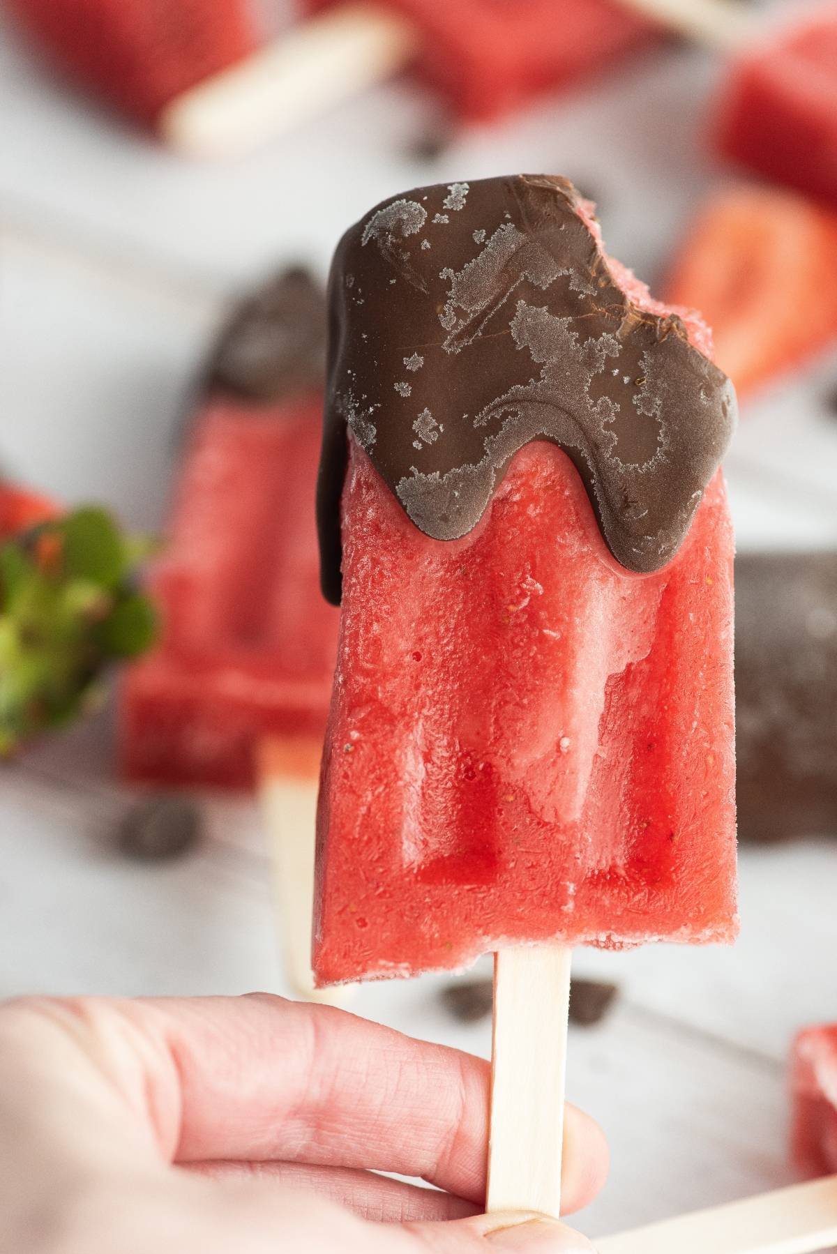 A hand holding a watermelon popsicle dipped in chocolate, with a bite taken from the top—a delicious summer treat recipe perfect for cooling off.