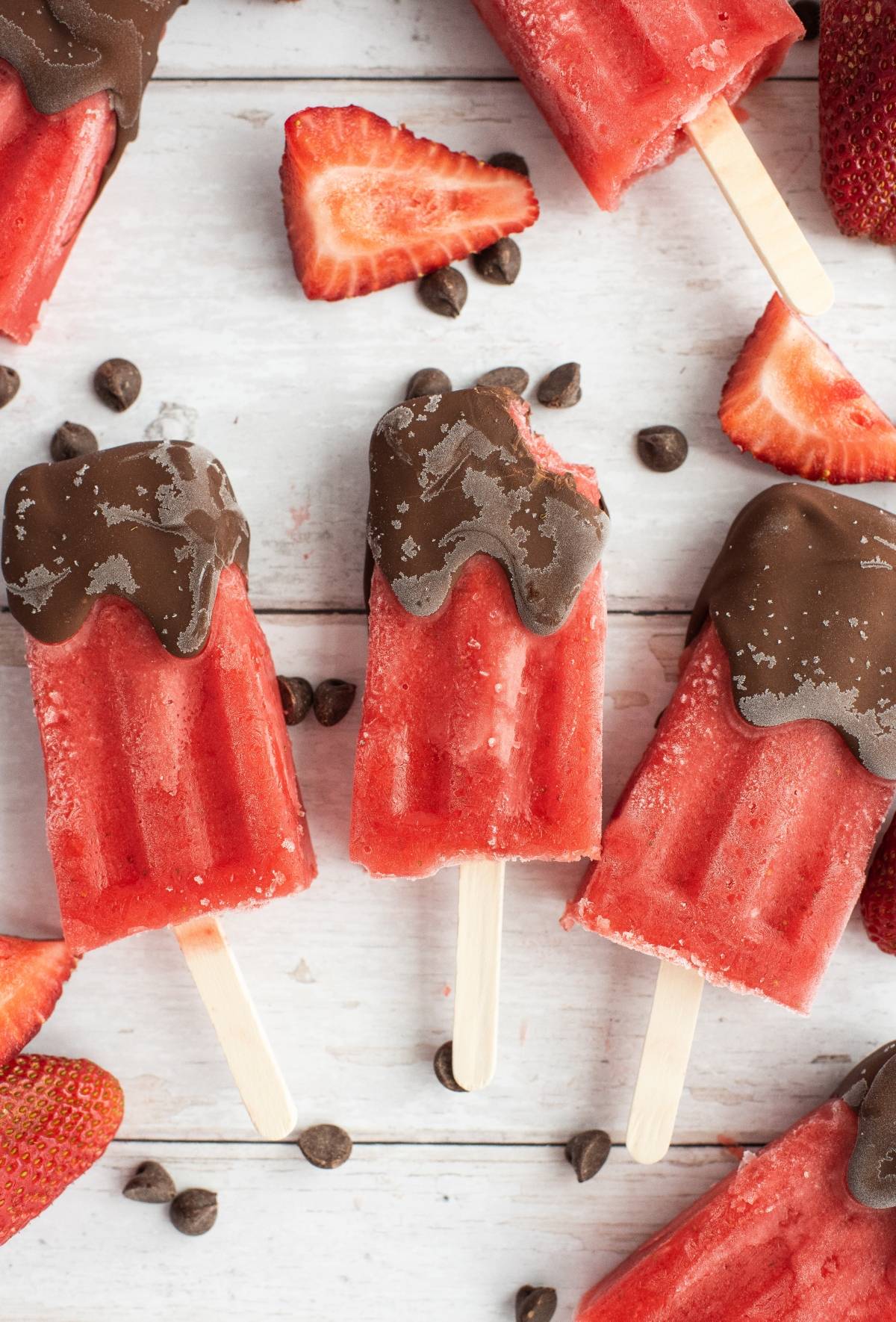 Three Chocolate Covered Strawberry Popsicles dipped in rich chocolate, surrounded by sliced strawberries and chocolate chips on a white surface. Try this dipped fruit popsicle recipe for a decadent chocolate covered fruit bar treat!.