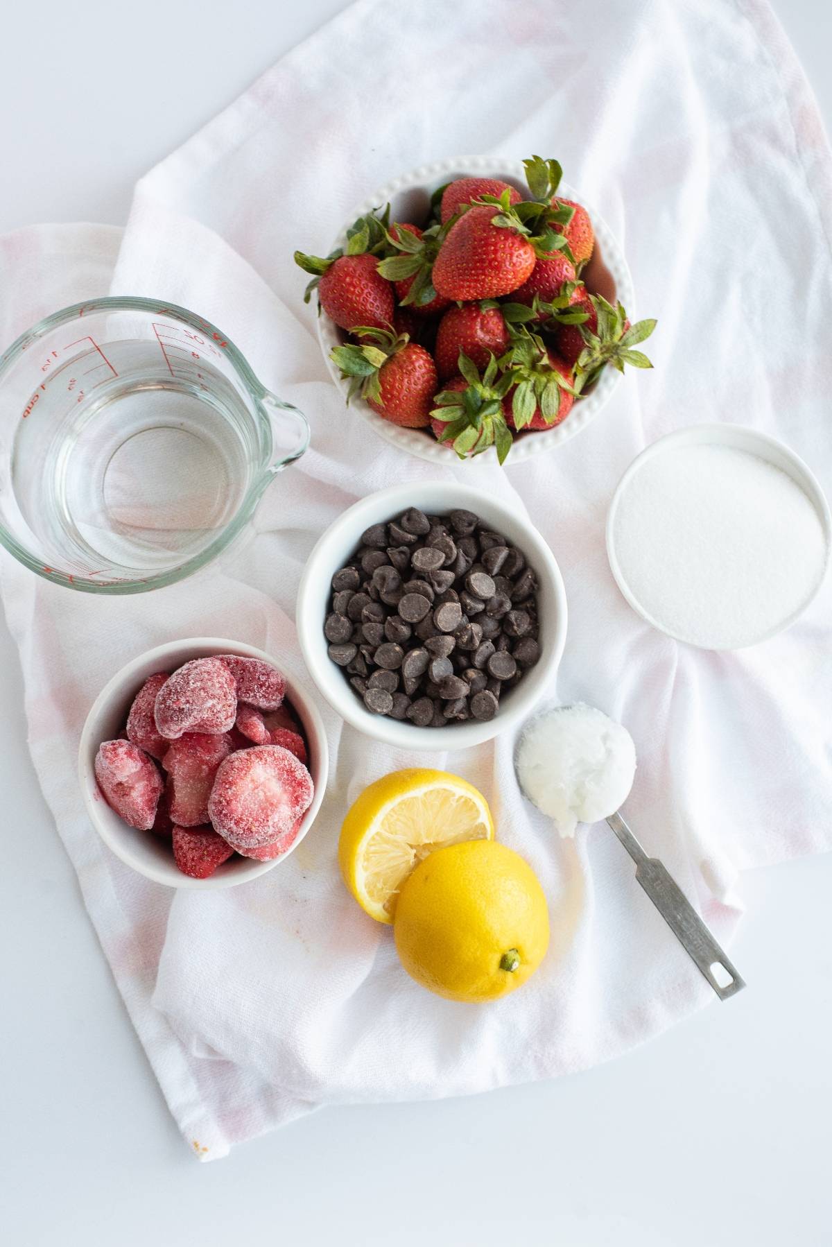 Strawberries, chocolate chips, lemon, frozen strawberries, sugar, and water rest on a white cloth—perfect ingredients for easy homemade popsicles or chocolate covered strawberry popsicles.