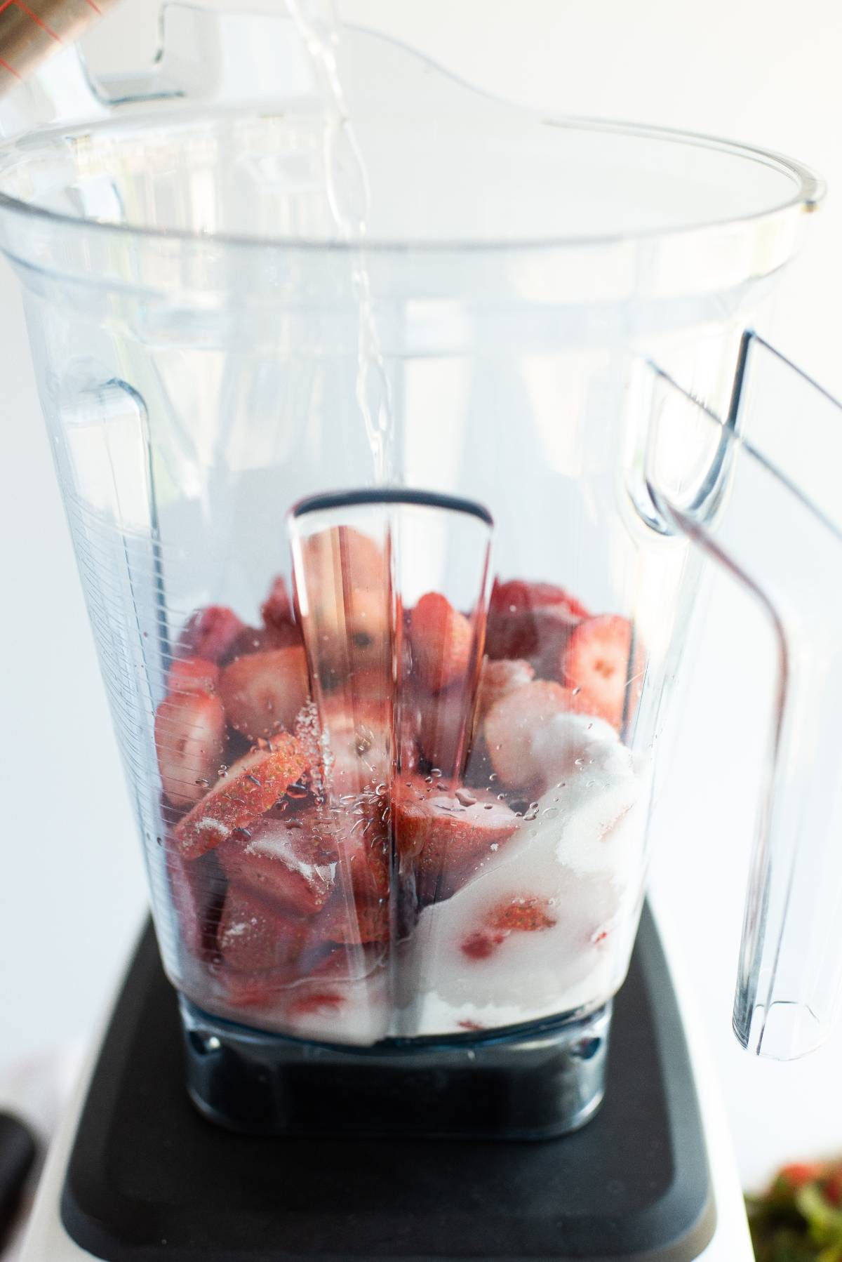 Sliced strawberries, sugar, and ice in a blender with water being poured in—perfect for making a refreshing chocolate covered strawberry popsicles summer treat recipe.