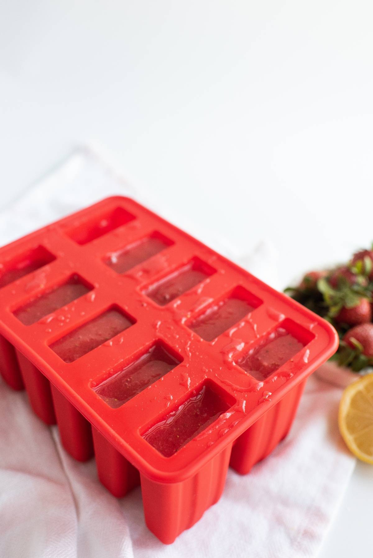 Red silicone popsicle mold filled with liquid sits on a white cloth, surrounded by strawberries and lemon—a perfect setup for an easy homemade popsicles summer treat recipe.