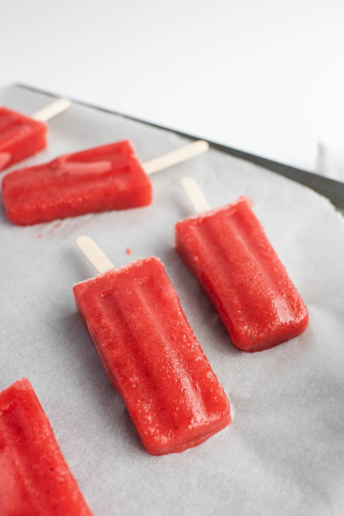 Three red easy homemade popsicles on parchment paper, with wooden sticks, rest on a white surface.