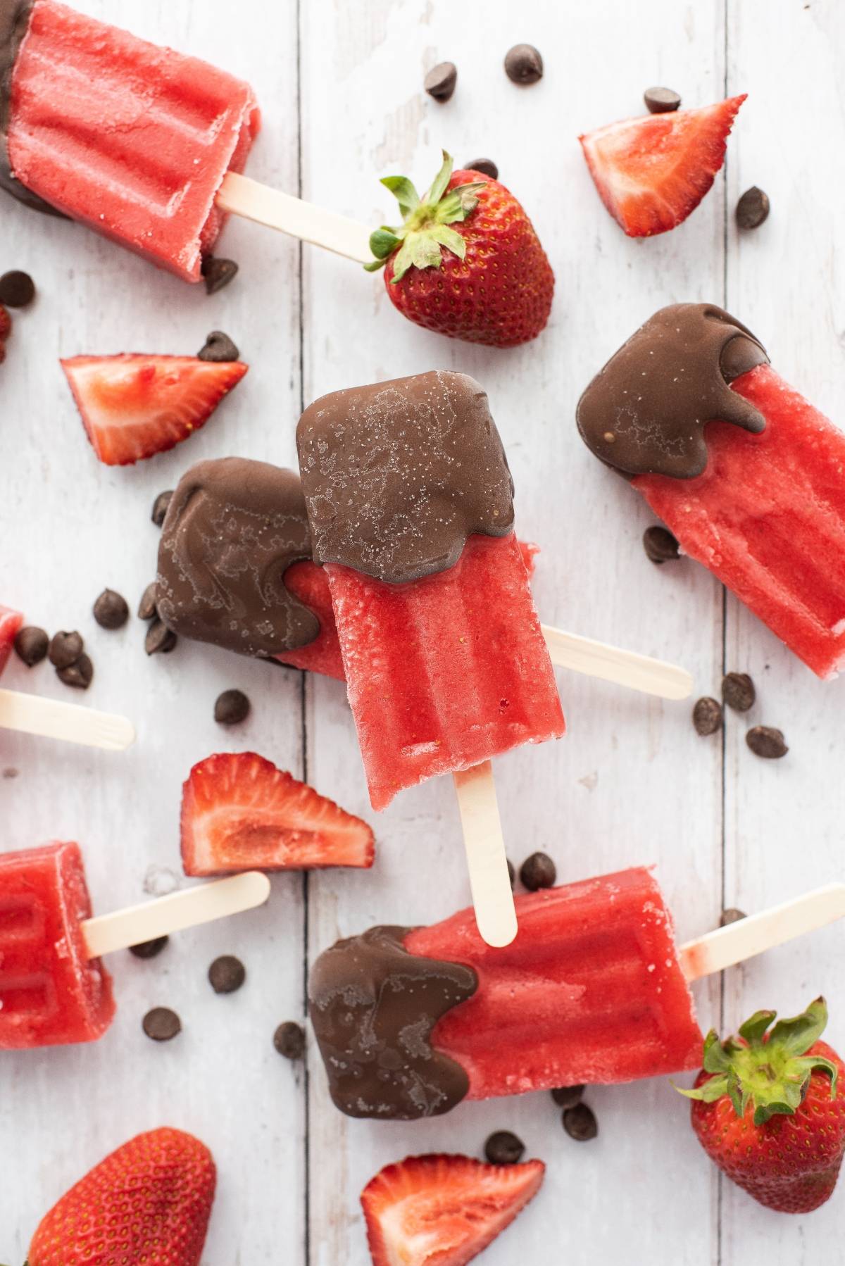 Strawberry popsicles dipped in chocolate, surrounded by fresh strawberries and chocolate chips—a perfect summer treat recipe for fans of chocolate covered fruit bars.