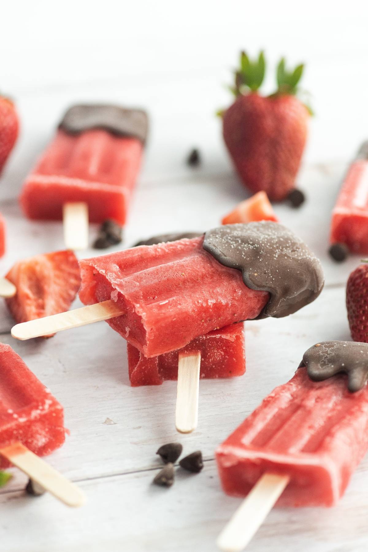 Easy homemade popsicles: Strawberry popsicles dipped in chocolate, topped with fresh strawberries and chocolate chips, make a delicious chocolate covered fruit bar treat on a white surface.