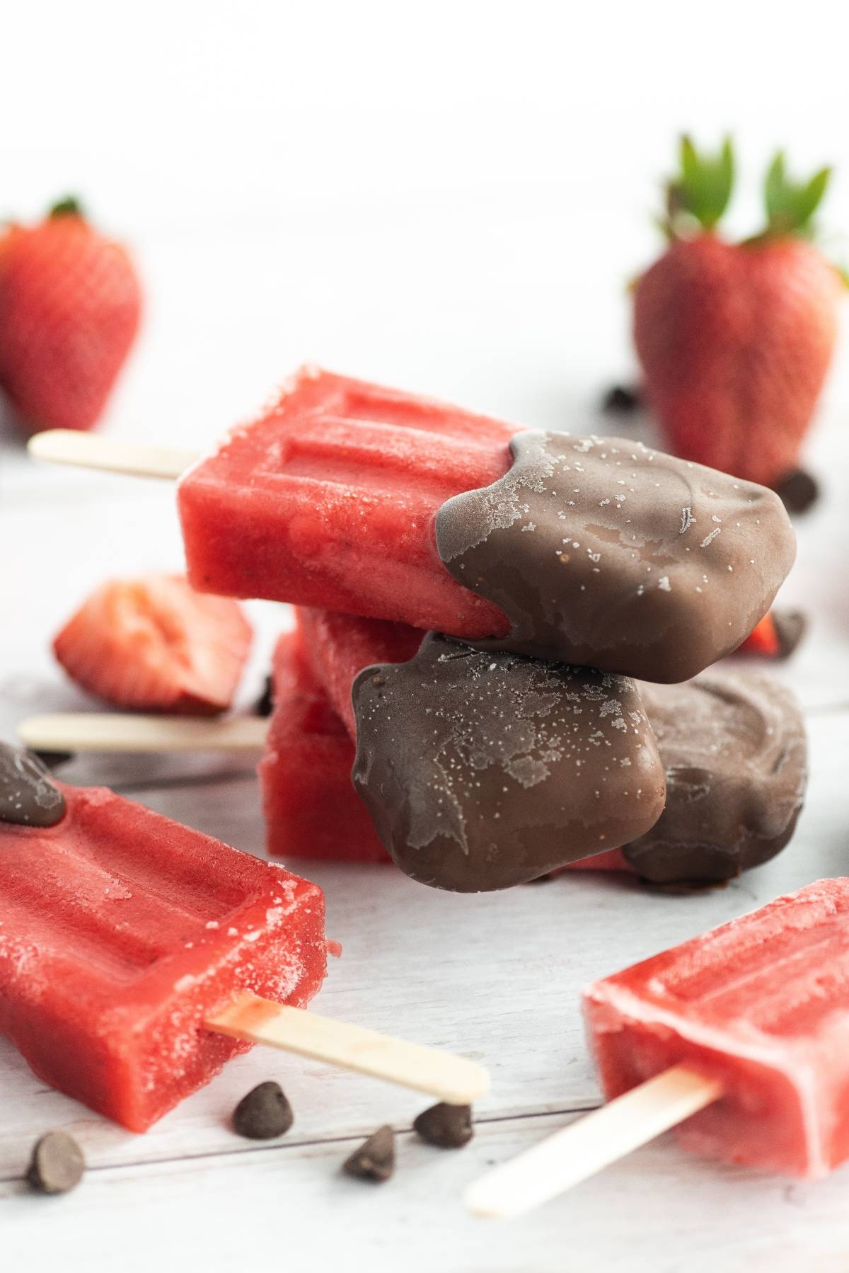 Chocolate Covered Strawberry Popsicles, some dipped in chocolate, are stacked with fresh strawberries and chocolate chips around—a perfect summer treat recipe.