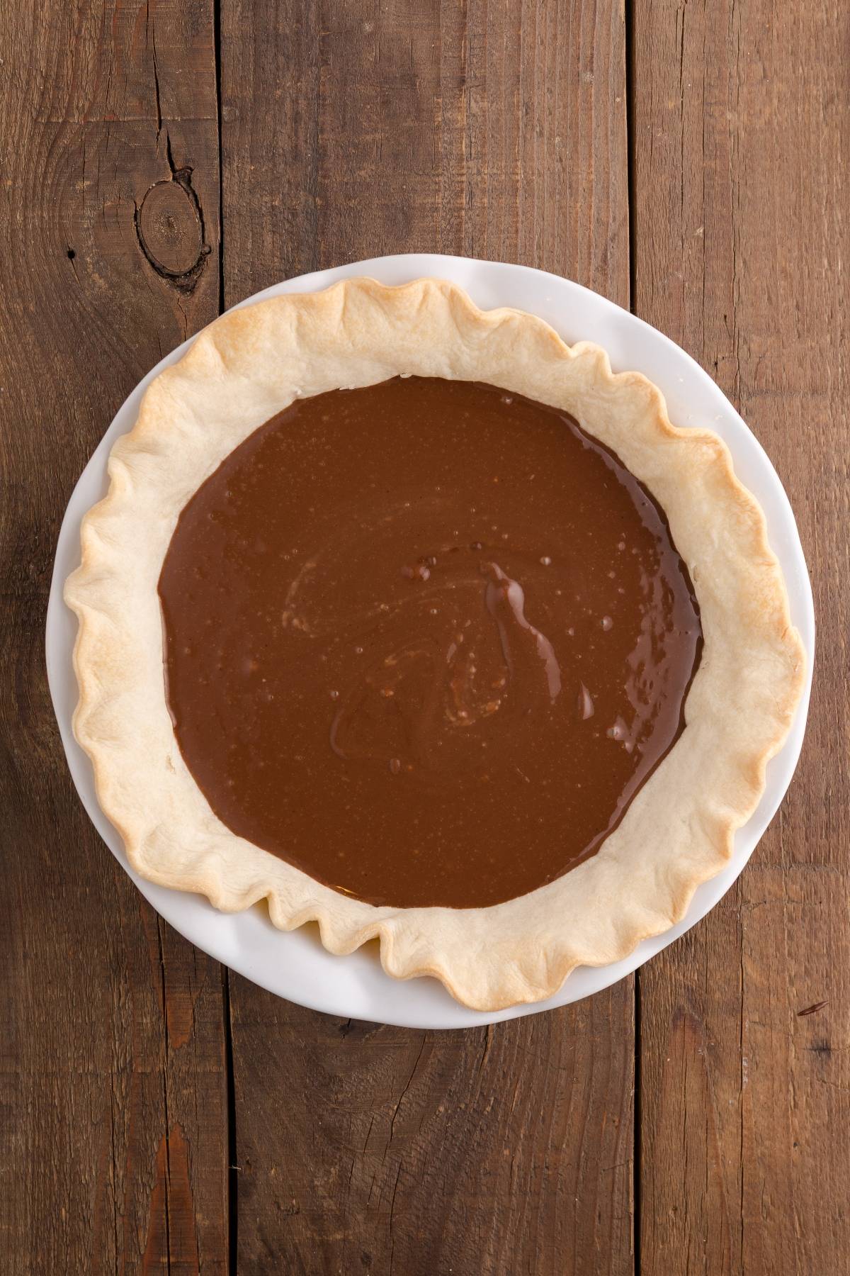Unbaked chocolate pie filling in a pie crust on a white plate, set on a wooden table.